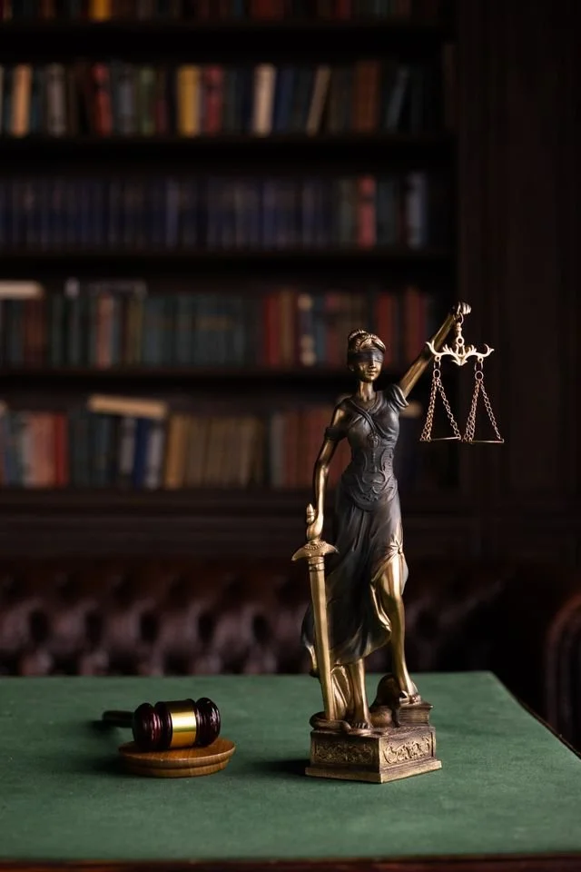 A dark-colored statue of Lady Justice holding balanced scales in one hand and a sword in the other, placed on a green table with a wooden gavel nearby, with blurred bookshelves in the background.