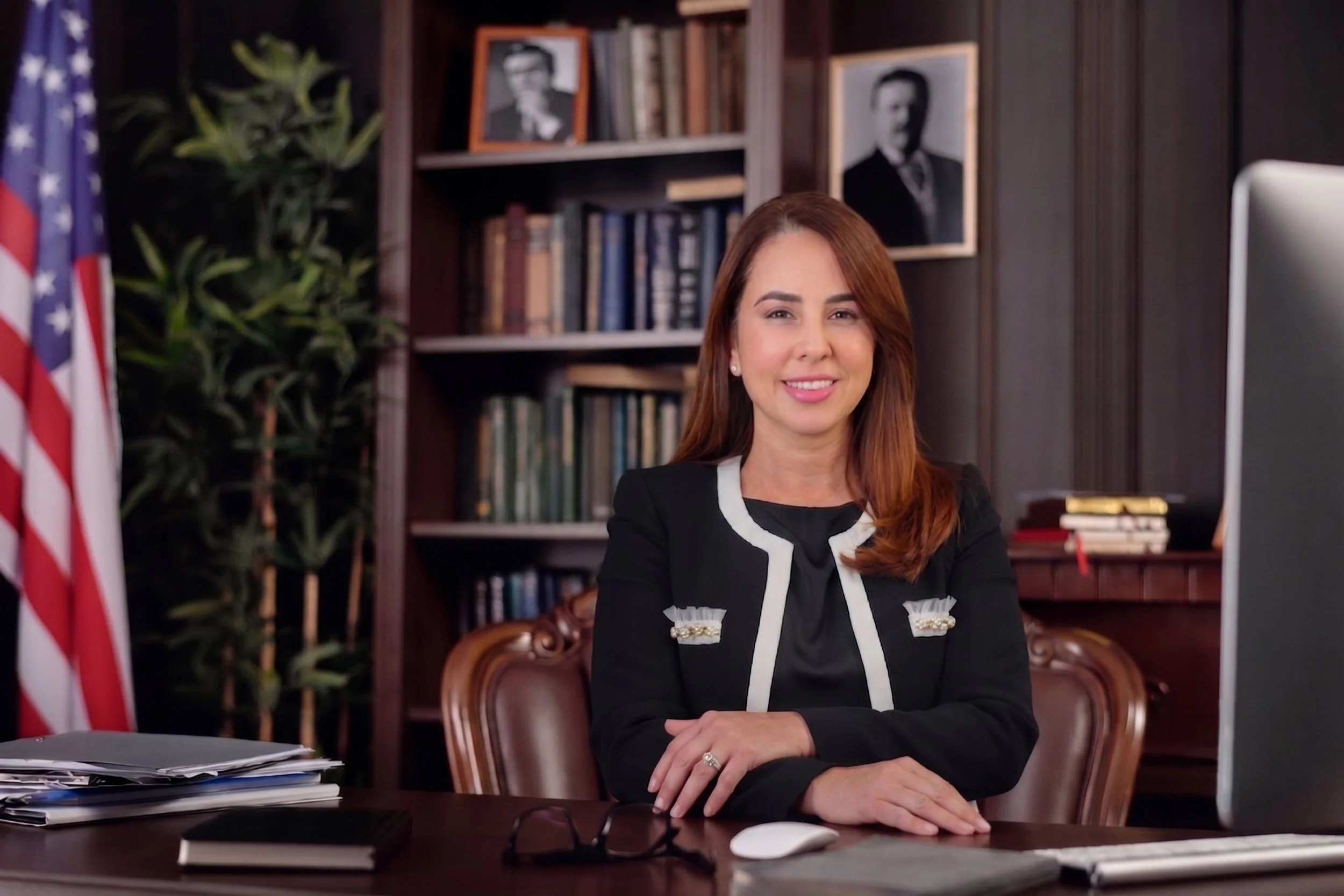 A woman with long red hair sitting at a desk with a computer monitor, papers, and a mouse in an office setting. She is smiling and wearing a black and white blazer. There are framed black-and-white portraits on the bookshelf behind her, a potted plant, and an American flag to the side.