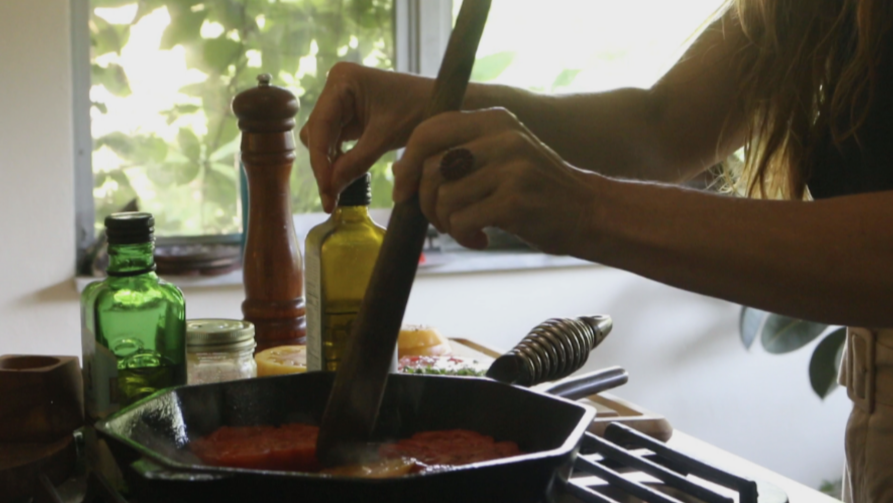 Chef's hands cooking at a stove with various condiments and bottles nearby, with a window showing greenery outside.