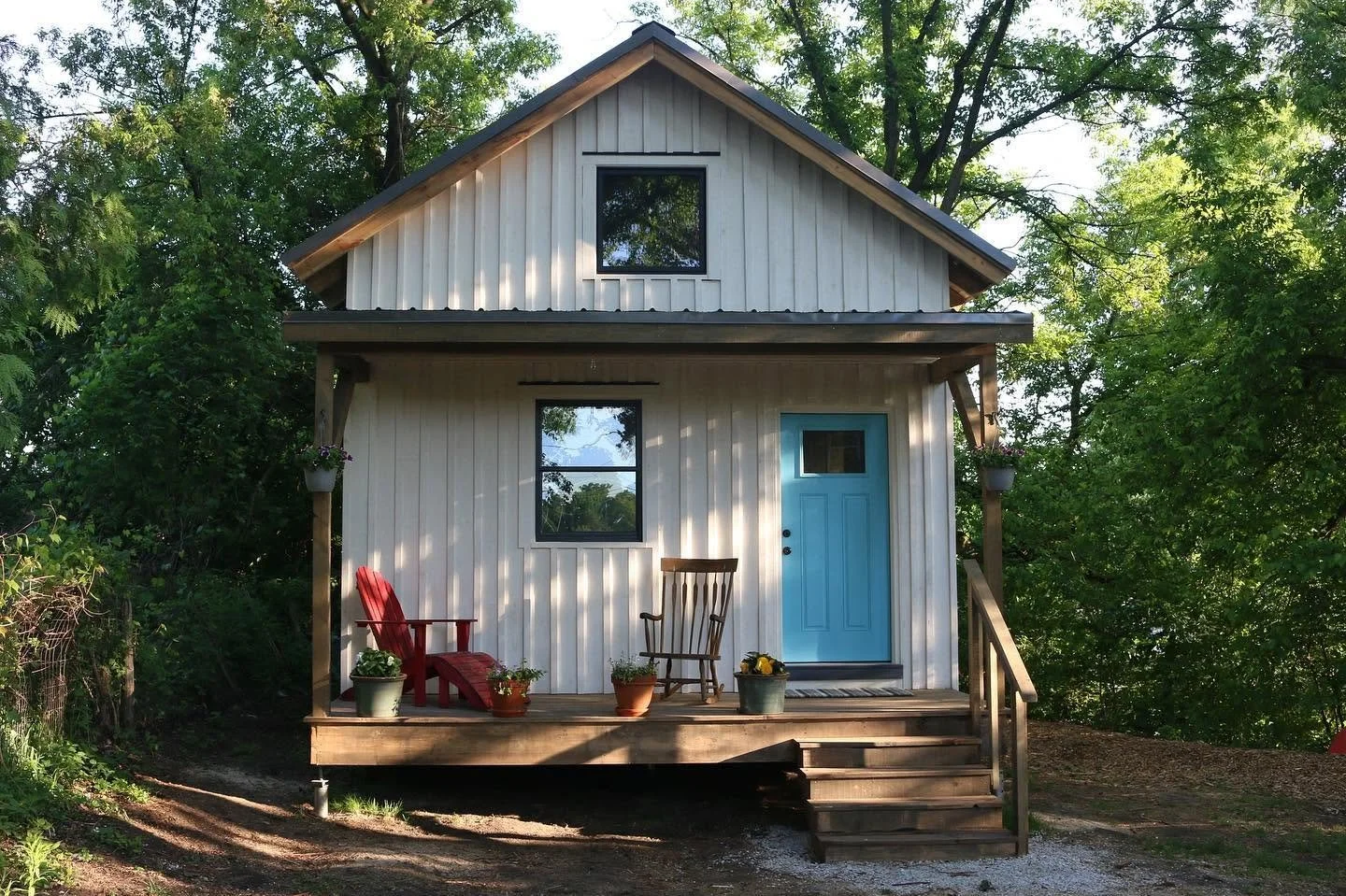 A small two-story white house with a light blue front door, wooden steps, and a porch with red and brown chairs, surrounded by green trees.