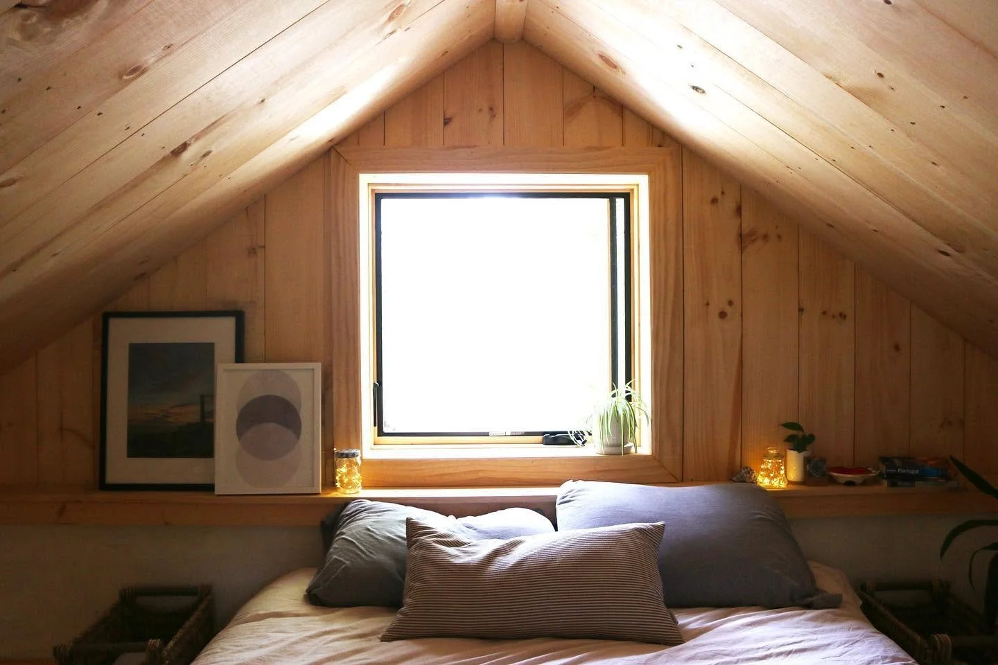 A cozy bedroom with a sloped wooden ceiling, a bed with gray and striped pillows, a window with a small plant on the sill, framed artwork, and decorative jars with lights on a shelf.