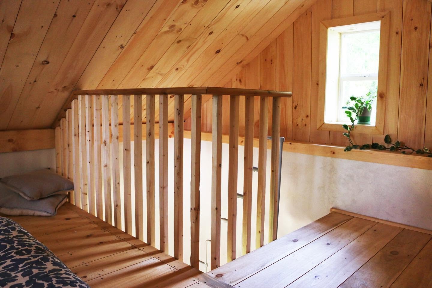 A cozy attic bedroom with wooden paneling on the ceiling and walls, a small window with a potted plant, and a wooden bed with a headboard and pillows.