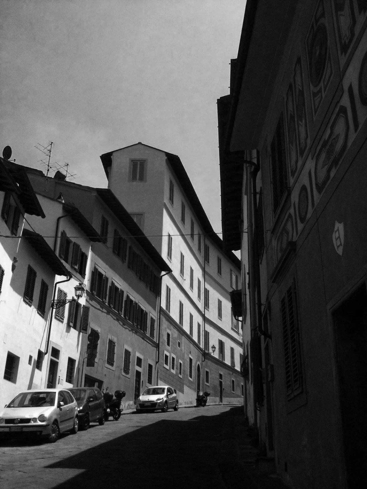 Black and white photo of narrow Florence, Italy street with cars parked along the curb, multi-story buildings with shutters and balconies, and a cloudy sky overhead.
