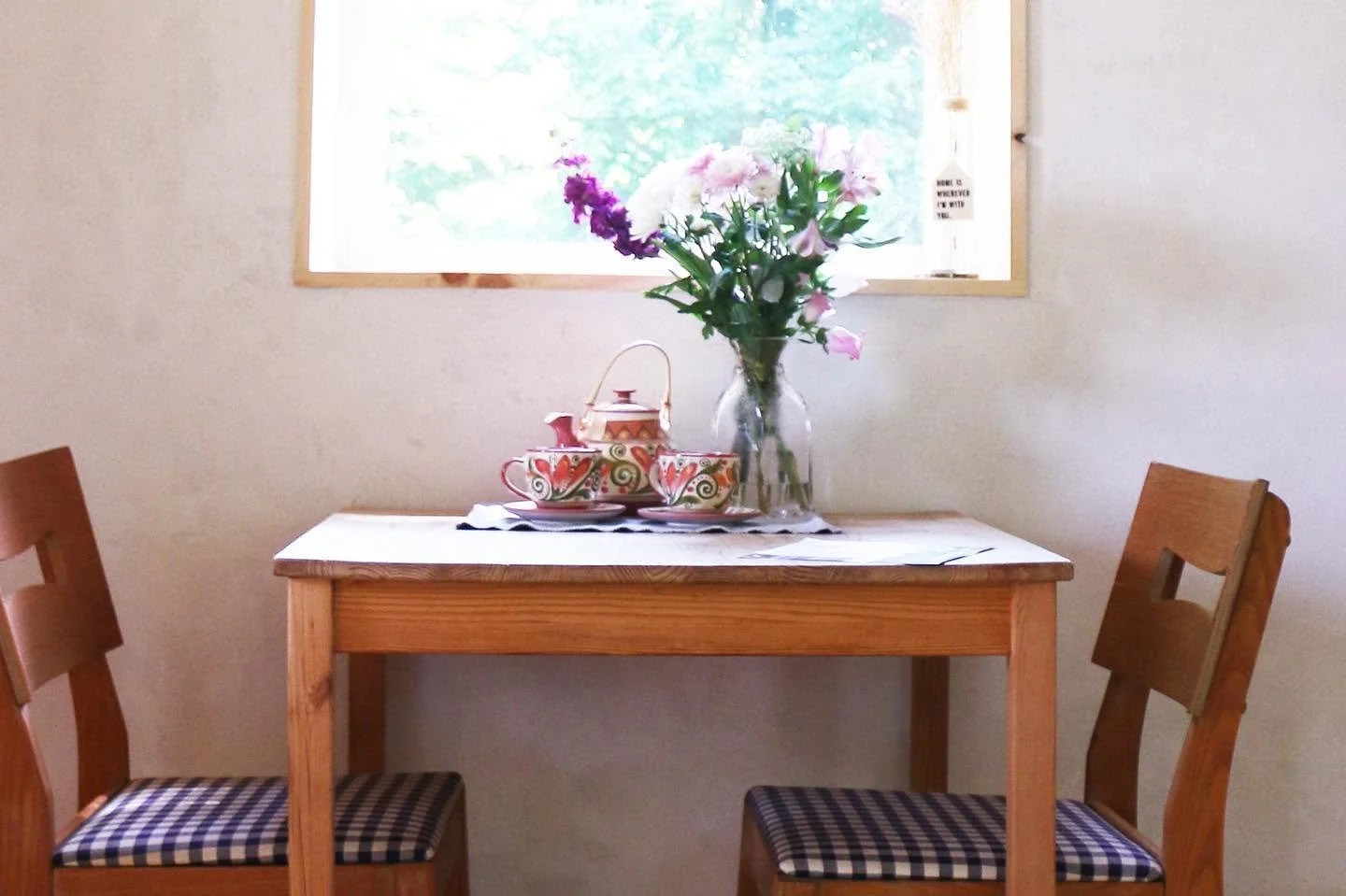 A wooden table with a vase of pink and purple flowers, a decorative tea set, and a place mat. Two wooden chairs with checkered seat cushions are on either side of the table, with a window in the background letting in natural light.