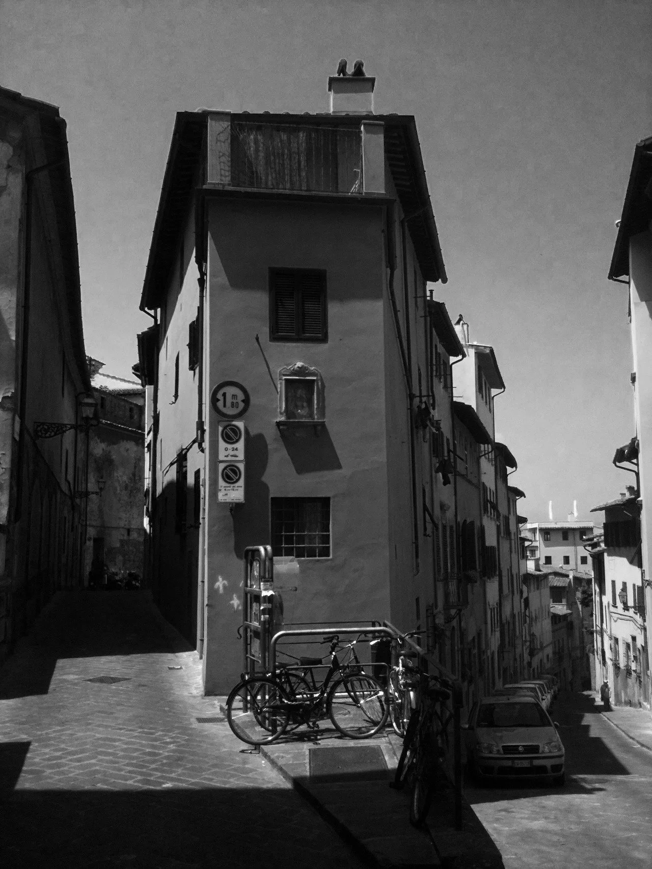 Black and white photograph of a narrow, multi-story Florence, Italy street with old buildings, parked bicycles, and cars, with street signs and shadows cast by the buildings.