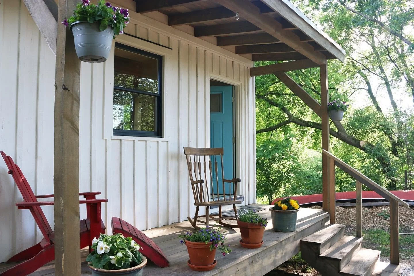 A small porch with potted flowers, a red chair, and a wooden rocking chair in front of a house with white vertical siding and a bright blue door. Hanging planters with purple flowers are on the porch roof. Trees and greenery surround the porch.