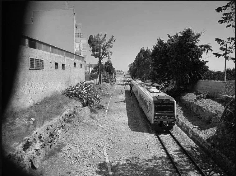 A black and white photo of an outdoor train track with a train approaching from the background, on the right side of the image. There are trees and an urban building on the left side. Bari, Italy.