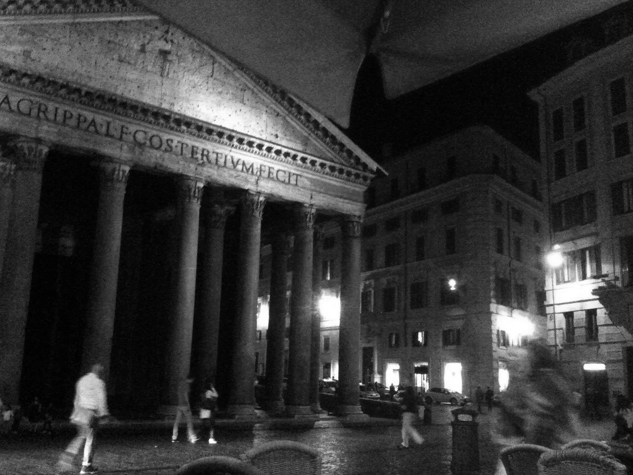 Nighttime scene of Pantheon in Rome, Italy with people walking in the foreground, illuminated by streetlights, with historic buildings surrounding it.