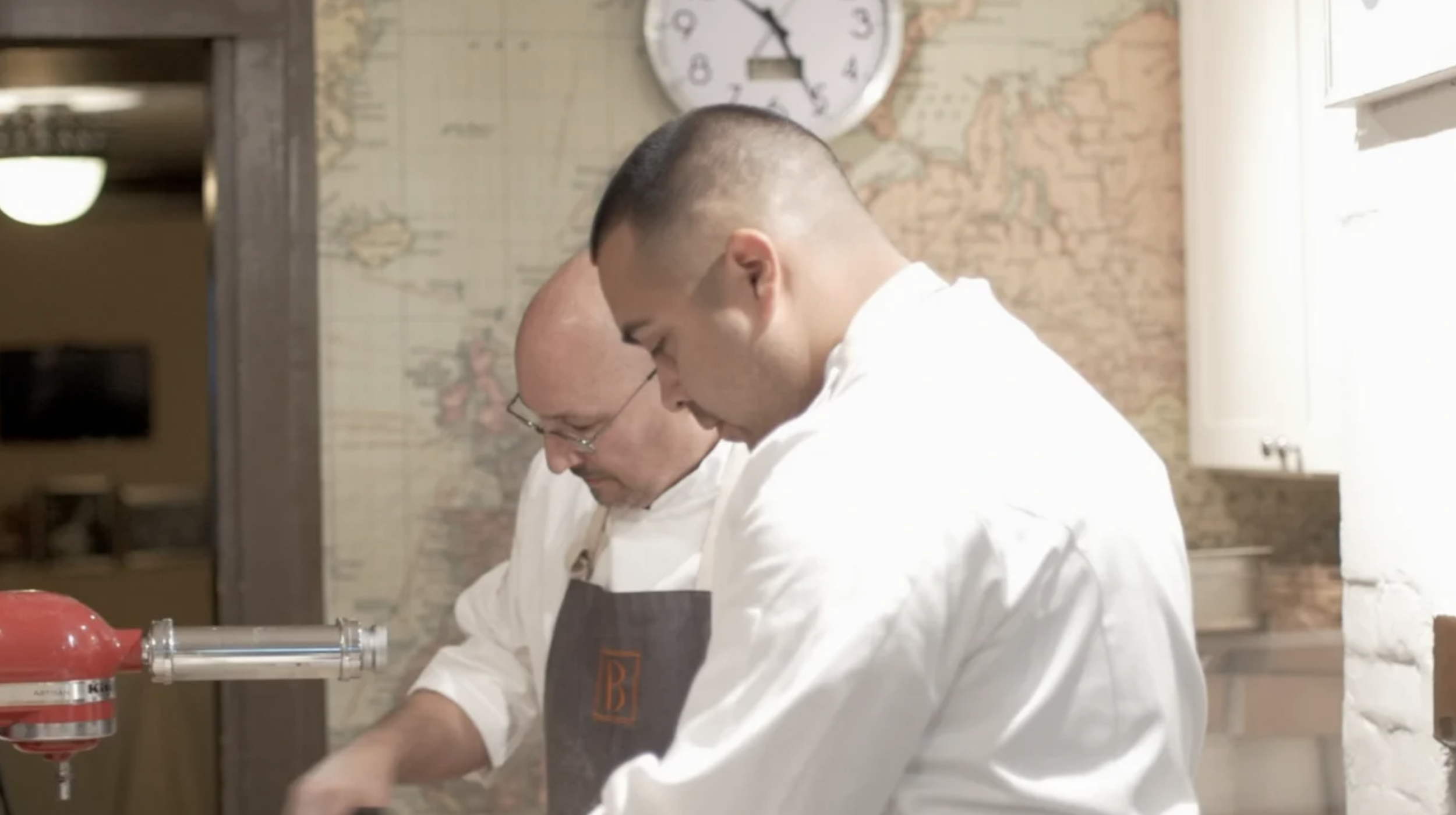 Two chefs in white chef aprons working together in the James Beard House kitchen, with a wall map and clock in the background.