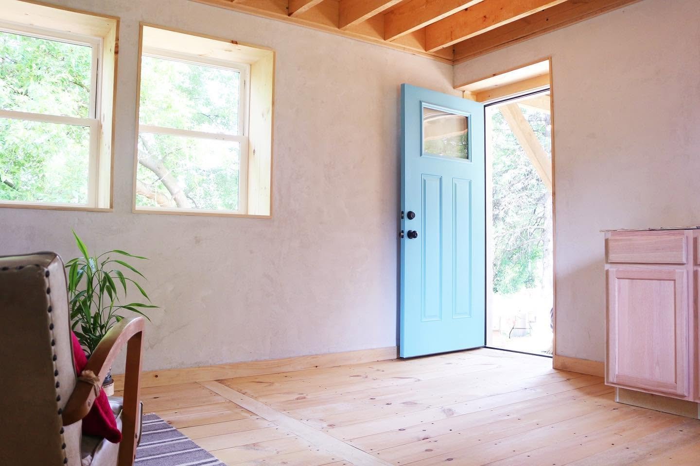 Bright room with light wooden flooring, two windows, an open light blue door, a pink cabinet, and a green plant.