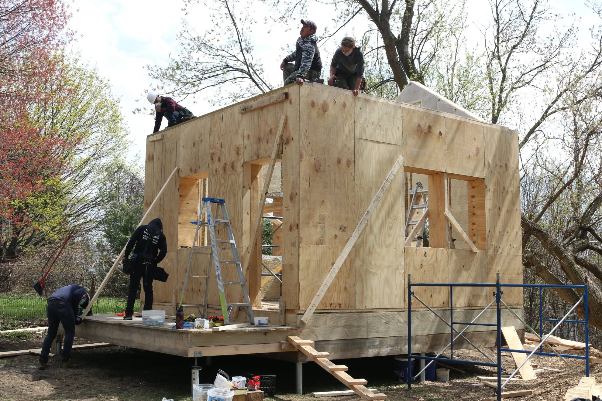 People working on constructing a wooden house in an outdoor area, with ladders and tools around, and trees in the background.