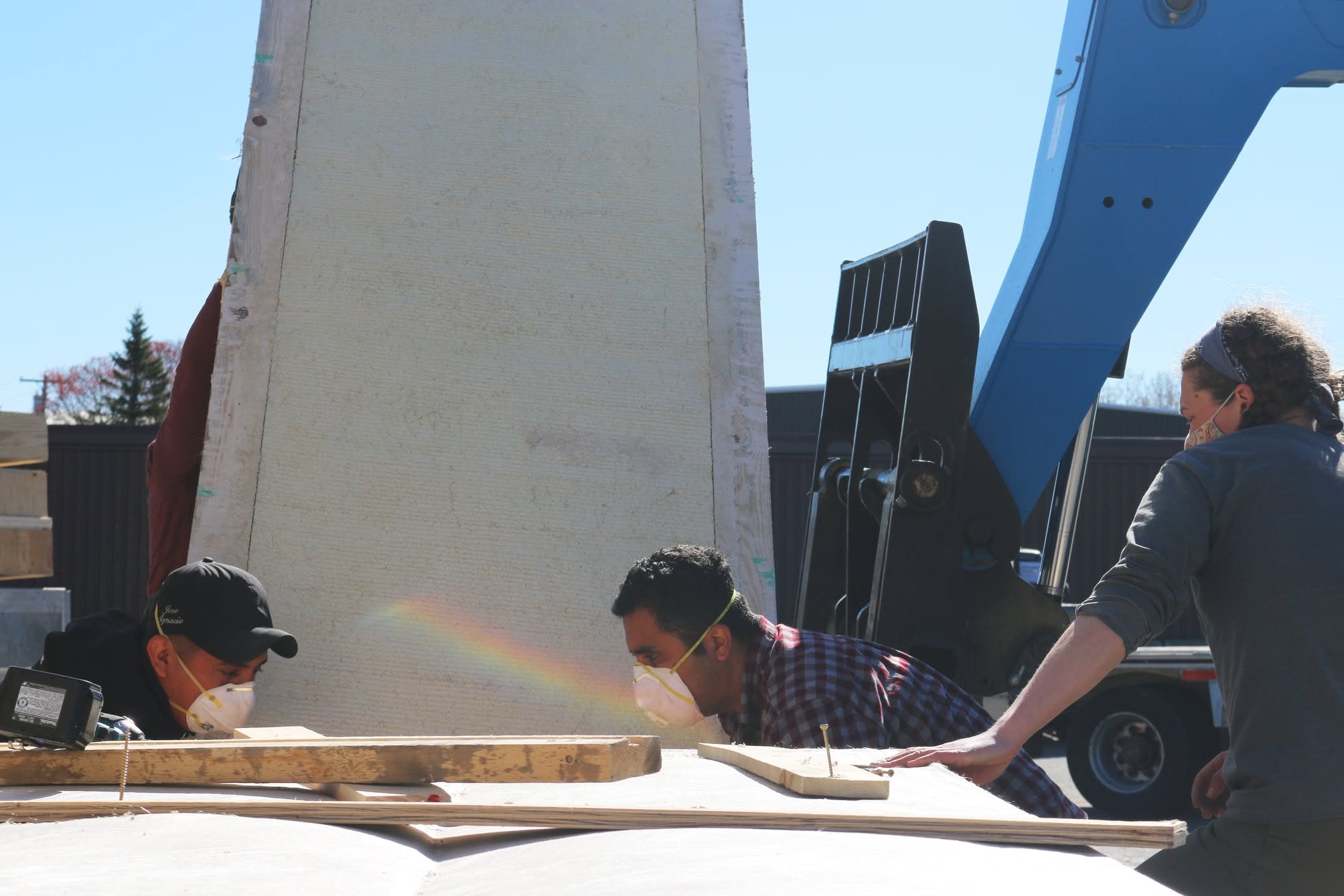 A group of people working together outdoors on a woodworking project. They are wearing masks and safety glasses, with tools and wood pieces on the table. A large piece of foam board and a piece of heavy machinery are visible in the background.