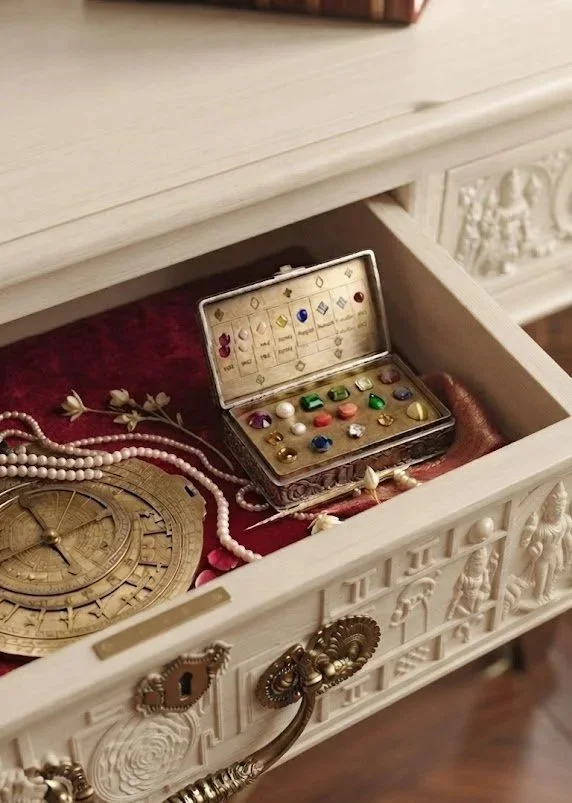 An open drawer containing vintage jewelry, beads, a clock, and a small jewelry box with colorful gems.