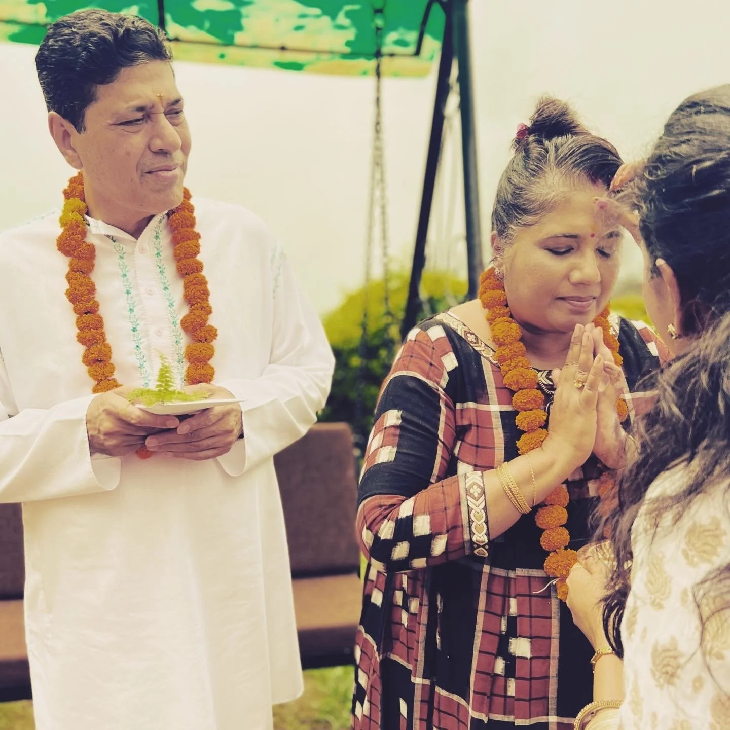 An Indian wedding ceremony with three women and one man, all wearing marigold flower garlands. The woman in the center is performing a traditional prayer with folded hands, while the man holding a plate stands nearby, smiling. The scene takes place outdoors, possibly under a canopy.