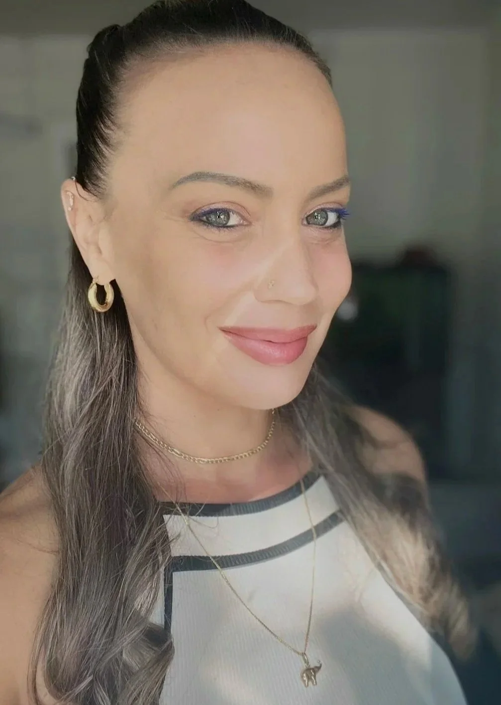Close-up of a smiling woman with long, wavy gray hair, wearing gold hoop earrings, layered gold necklaces, and a striped top.