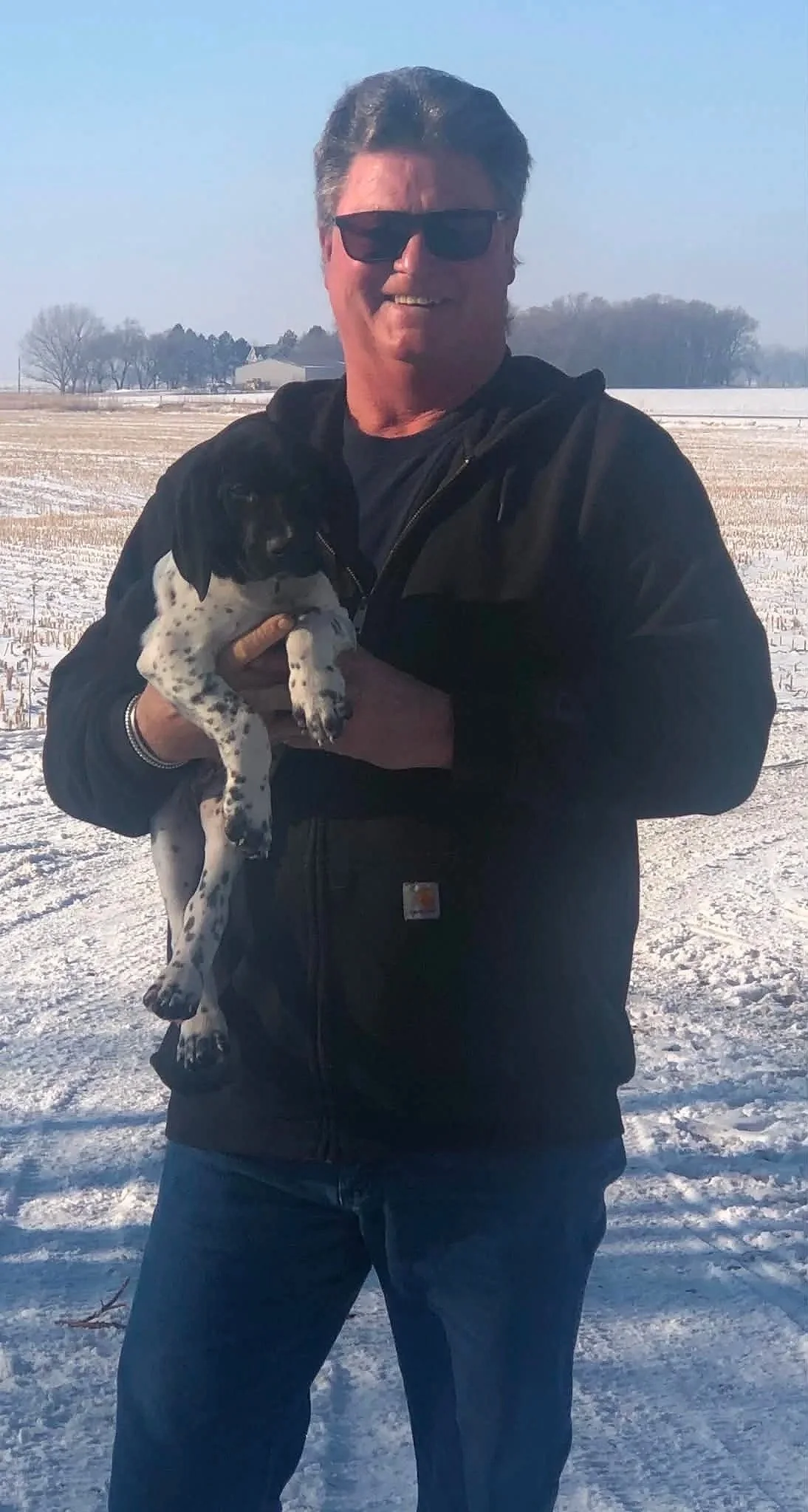 A man wearing sunglasses, a black jacket, and jeans standing outdoors in a snowy field, holding a small black and white puppy.