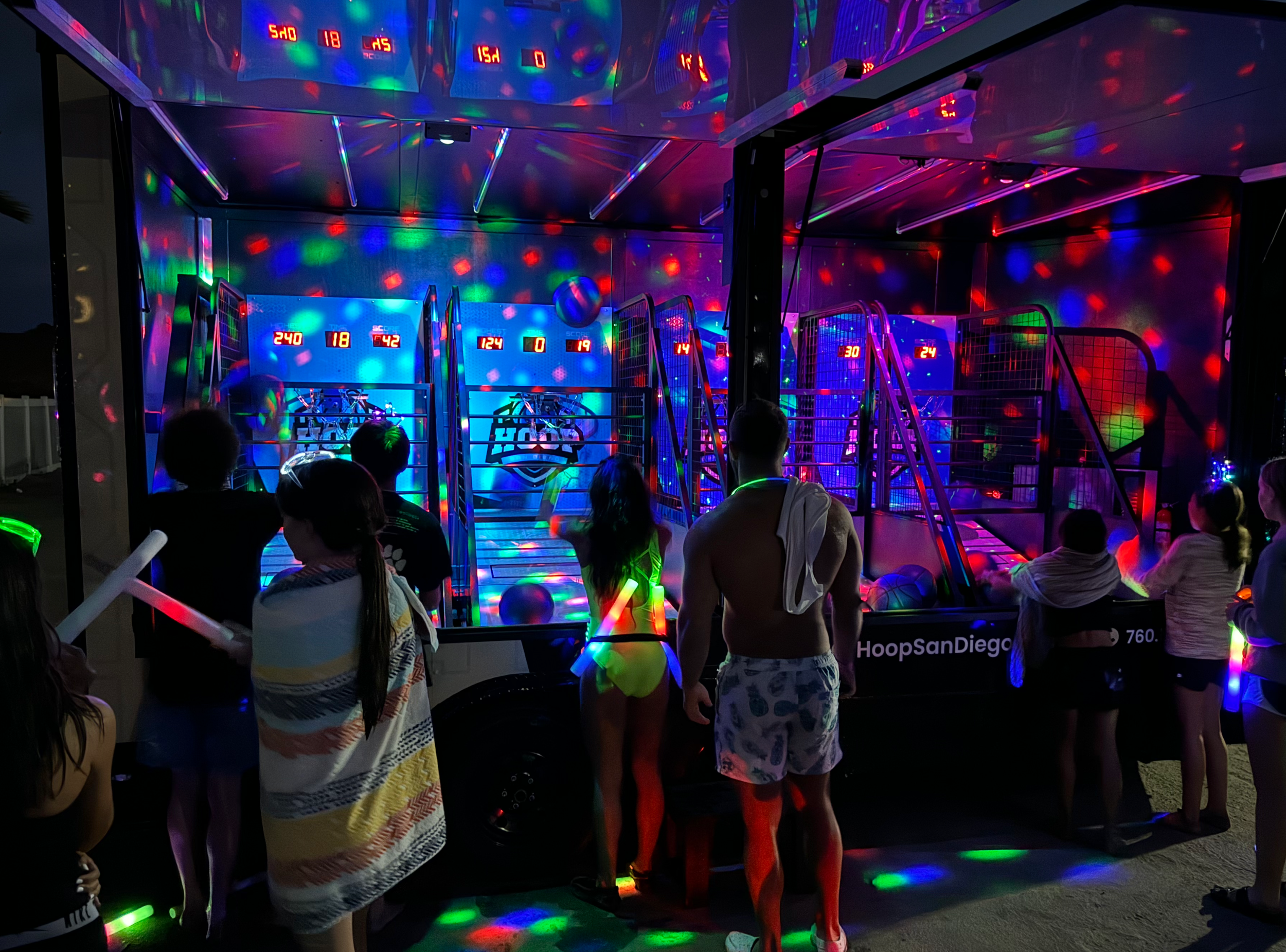 People standing in front of a colorful, illuminated indoor arcade game with digital score displays and red, green, and blue lights creating a vibrant atmosphere.
