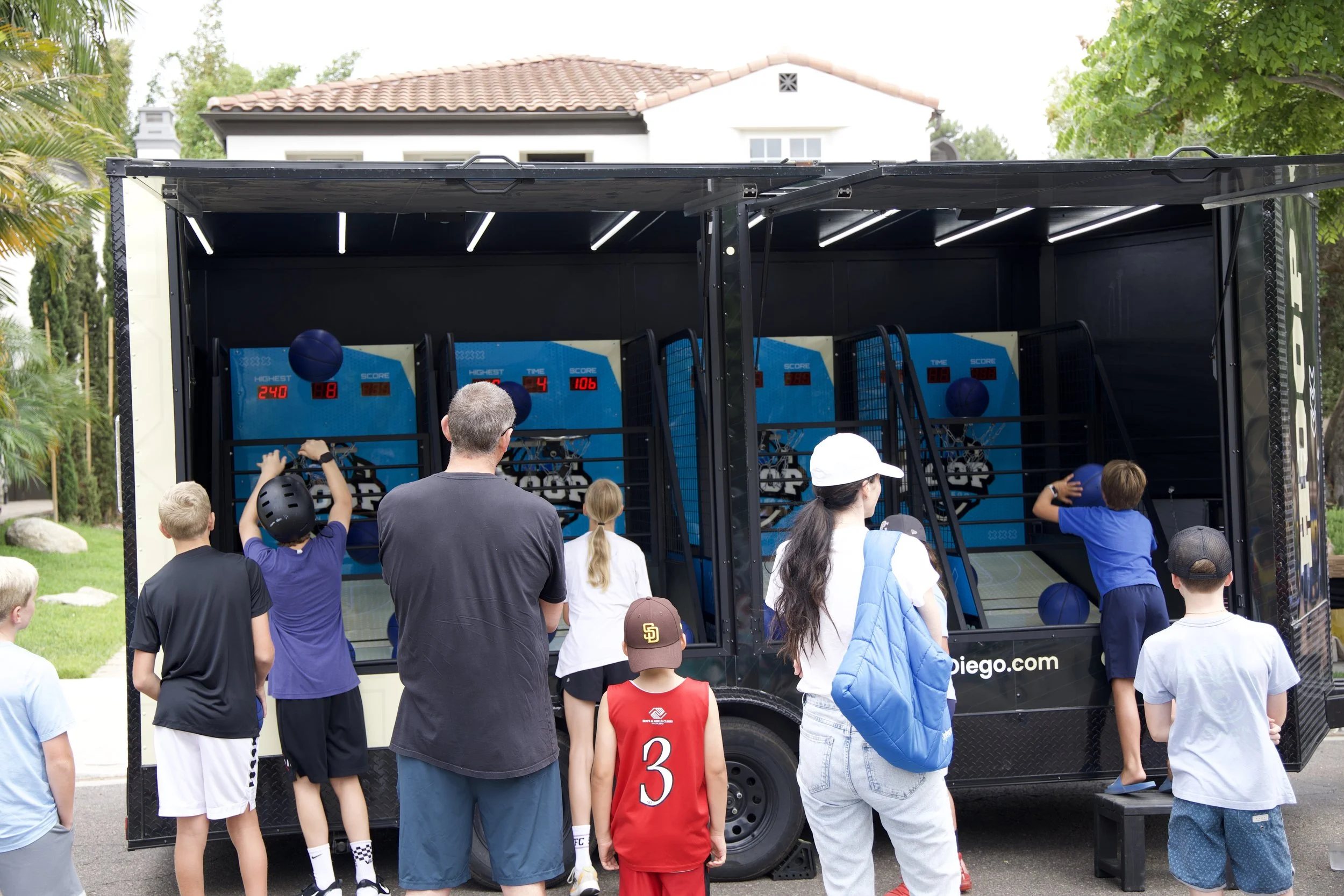 People gather outside a mobile basketball arcade truck, with children playing basketball games inside. The truck has LED lights and high-score displays. The background shows a house and trees.