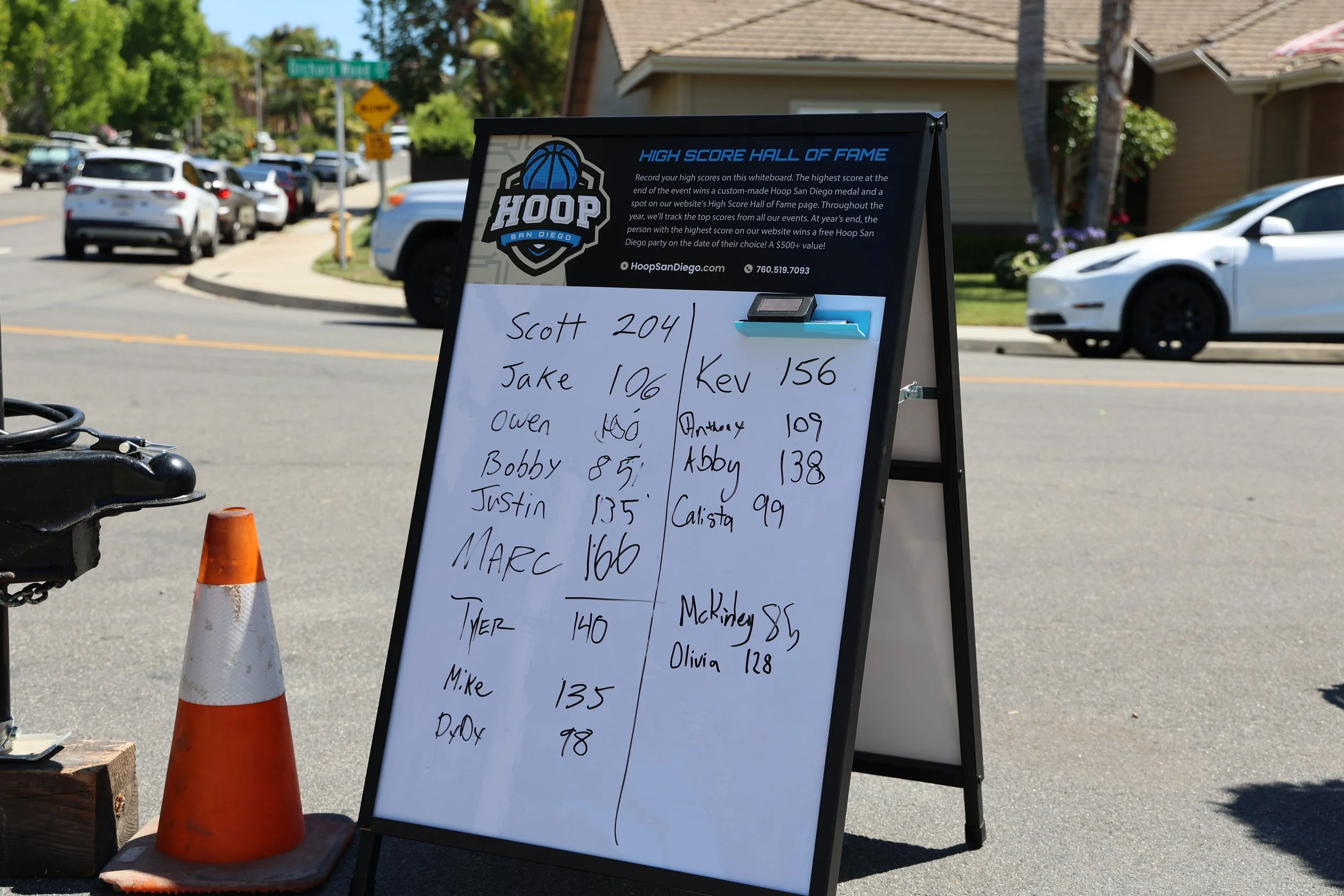 A black A-frame sidewalk signboard displaying high scores for a basketball game at Hoop San Diego. The board has a whiteboard with handwritten names and scores, and some branding and information about the event. An orange safety cone and a black object are in the foreground. The background shows parked cars along a residential street with houses, trees, and street signs.