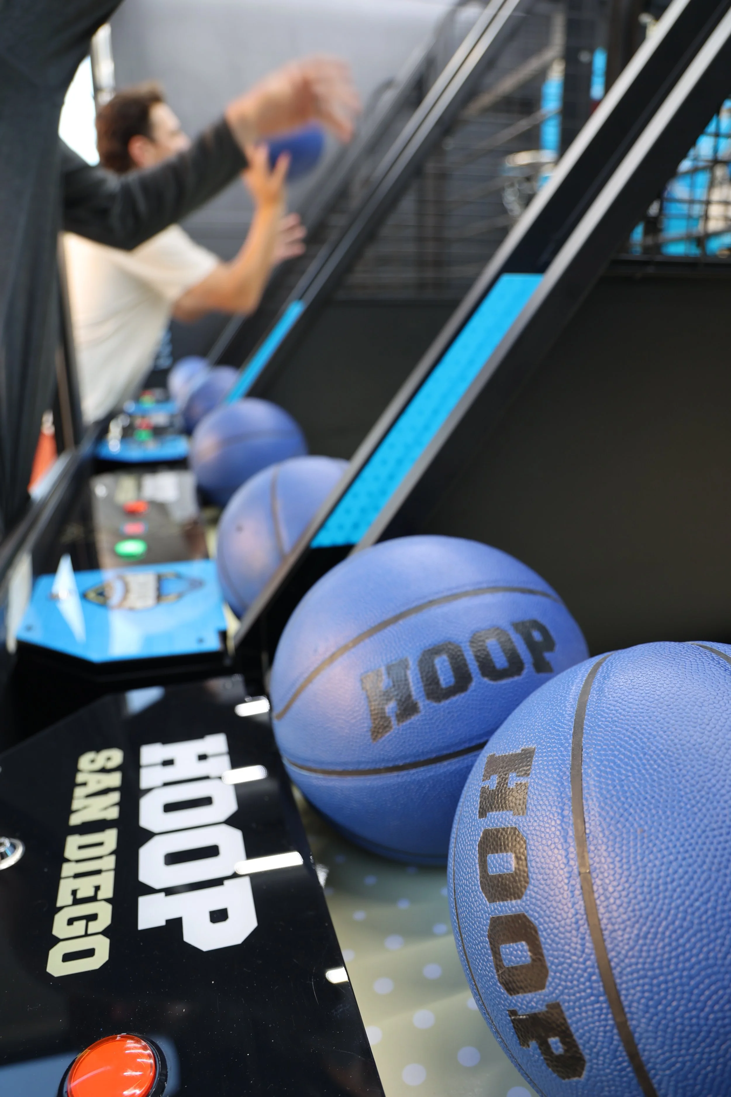 Several blue basketballs on a black gaming table with a red button, and two people in the background near a black staircase.