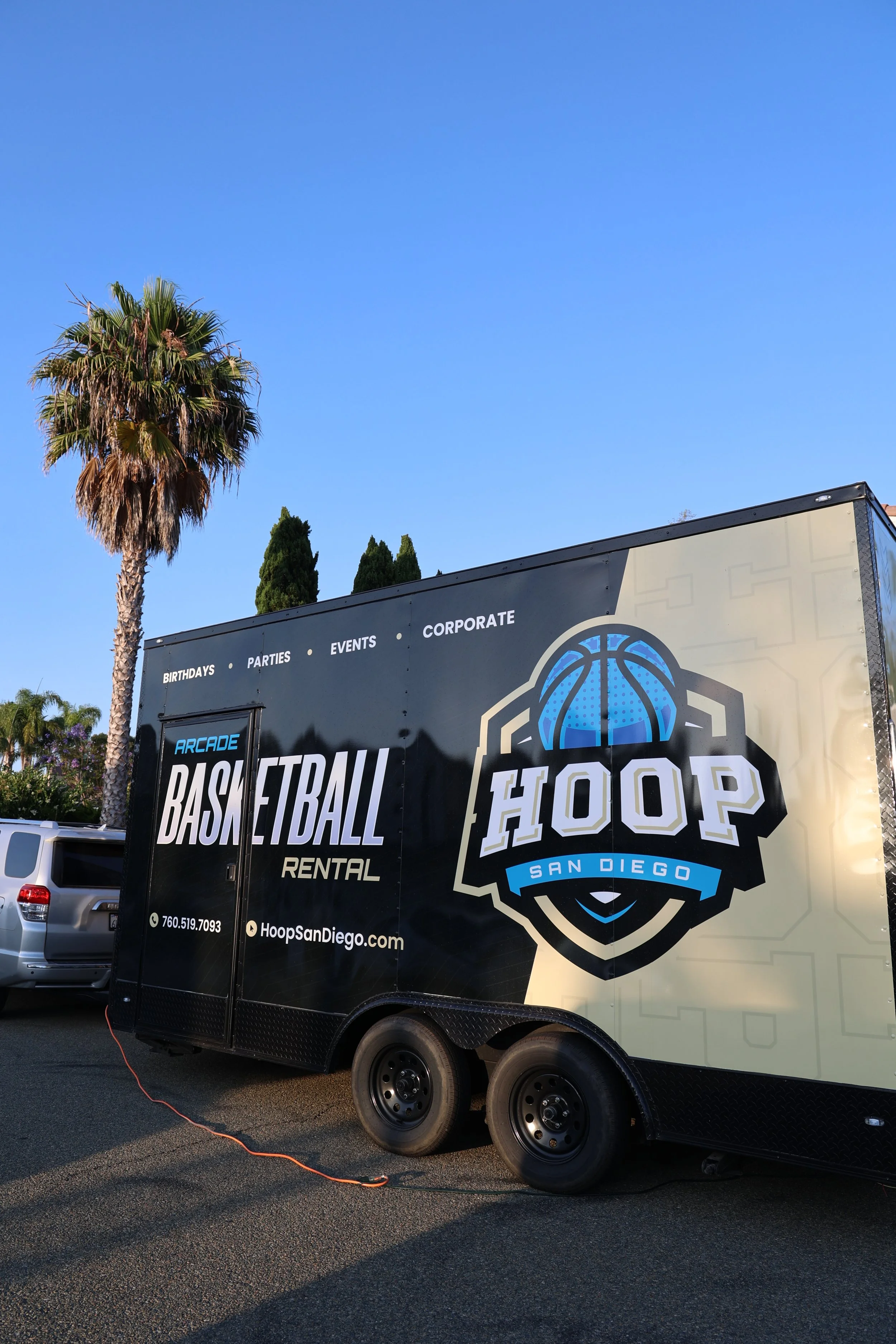 Black basketball Hoop Rental trailer in a parking lot with palm trees and blue sky in the background.