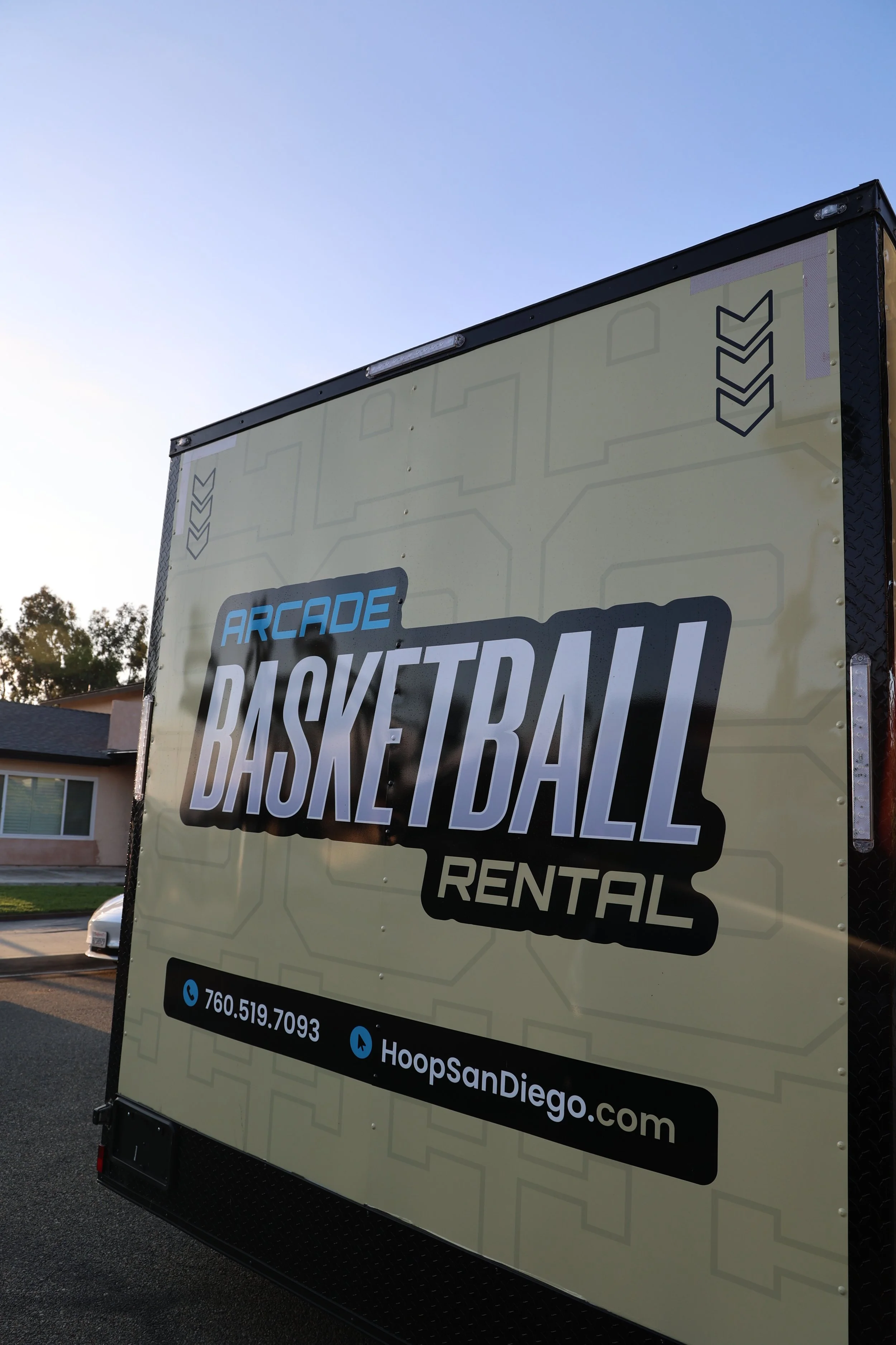 A mobile billboard promoting a rental service for arcade basketball, with contact details and a website, parked outside near residential buildings and trees under a clear sky.