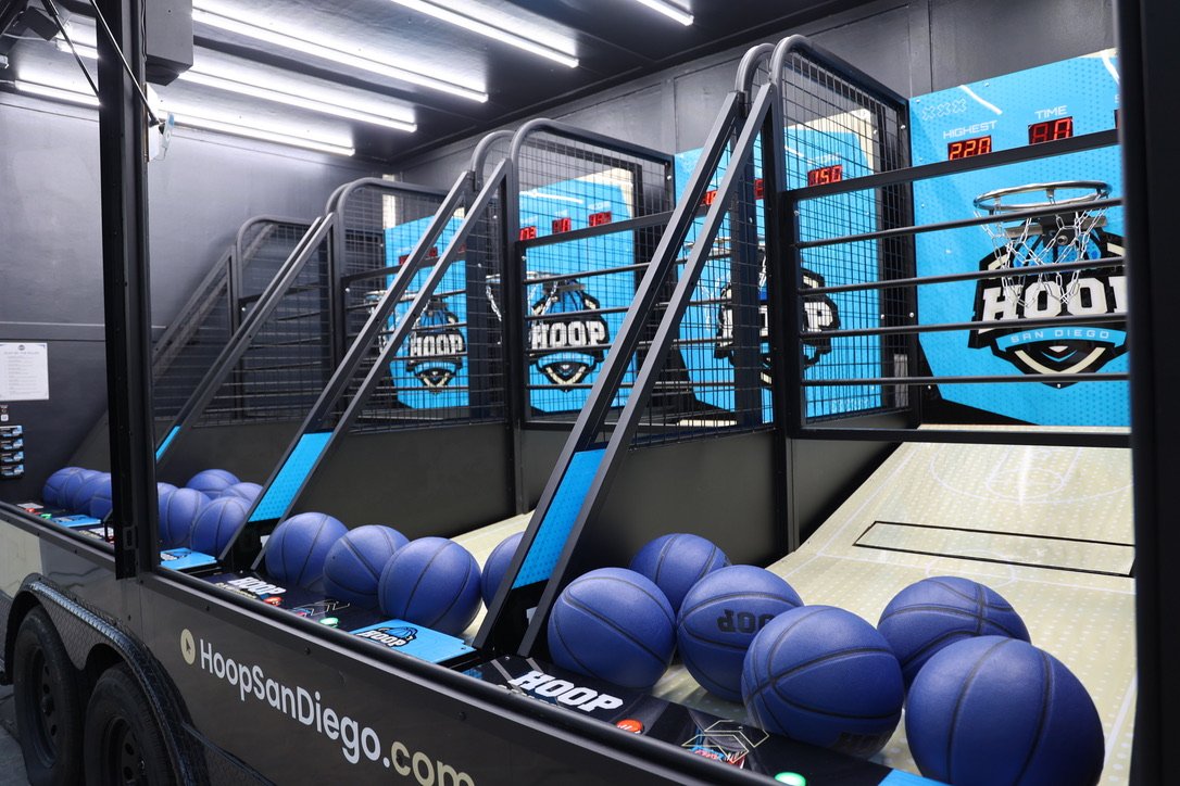 Indoor basketball arcade game with blue basketballs and digital scoreboard, branded 'Hoops San Diego'.