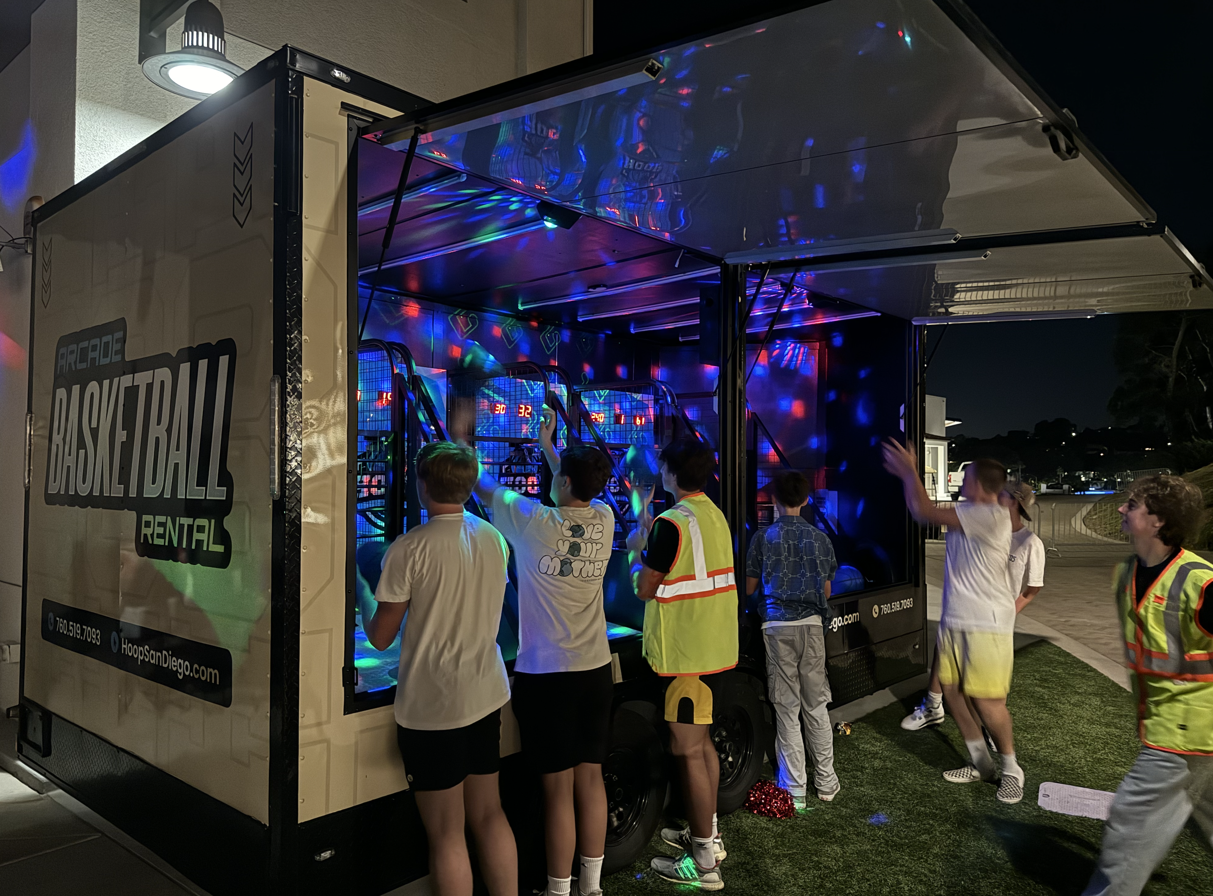 Kids playing a basketball arcade game inside a mobile gaming trailer with colorful lights at night.