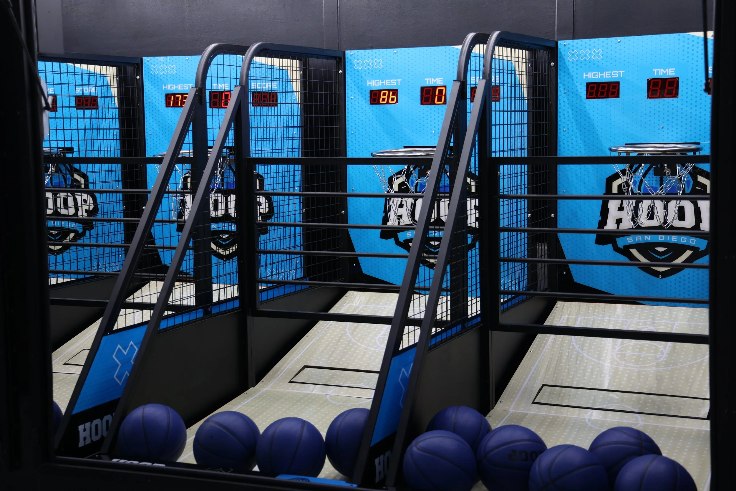 Indoor basketball shooting game with blue backboards, digital scoreboards, and black metal fencing, featuring basketballs lined up at the front.