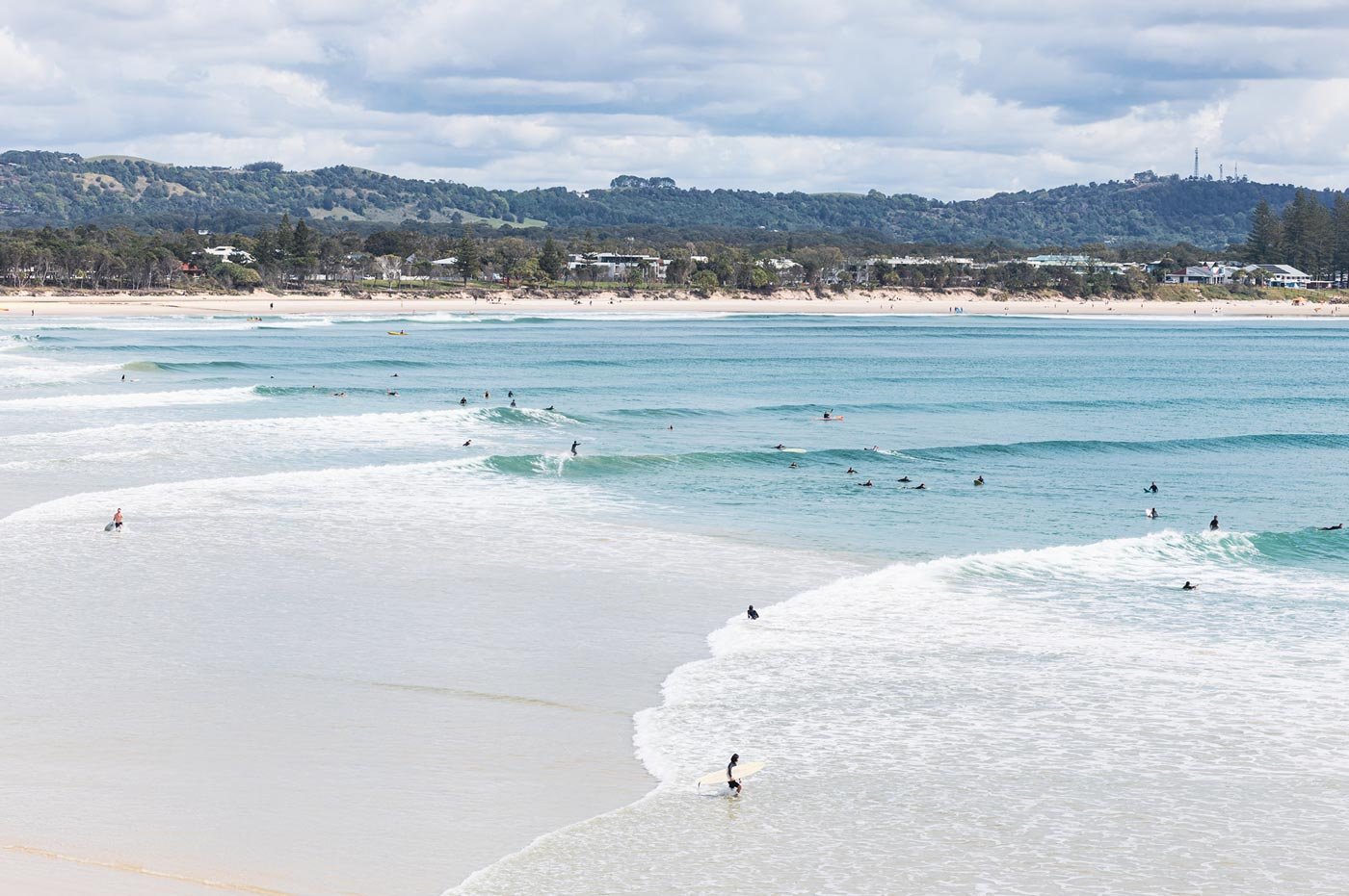 People surfing and swimming in the ocean at a sandy beach with green hills and houses in the background.