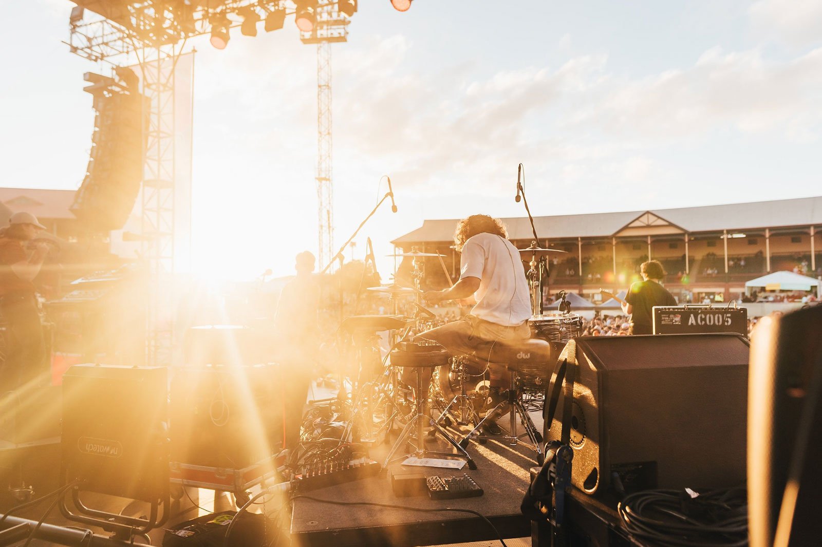 Musicians performing on an outdoor stage during sunset with crowd and festival infrastructure in the background.