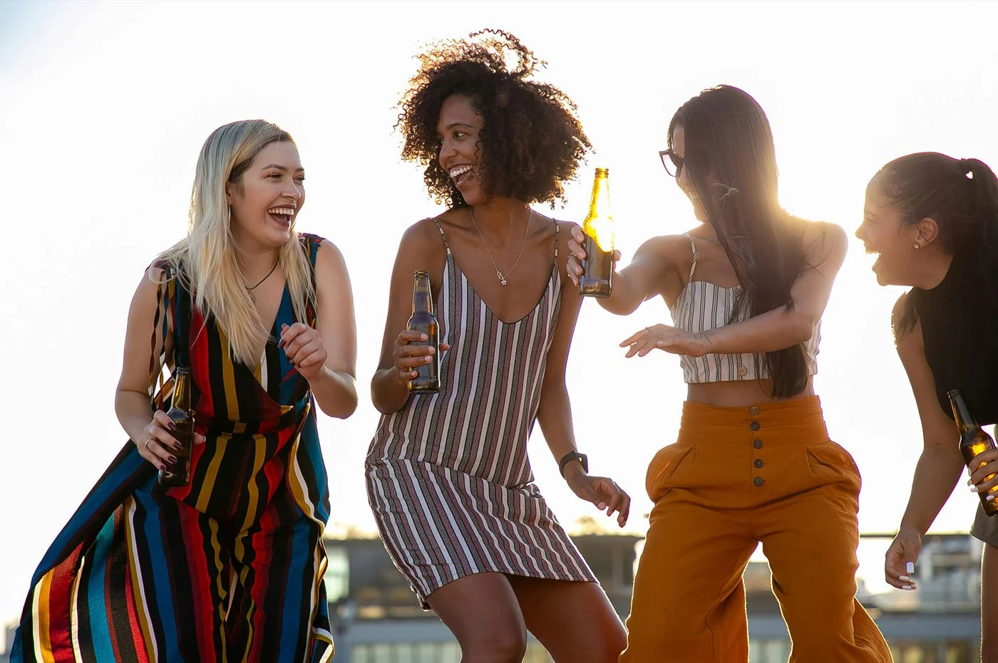 Five women enjoying drinks and dancing outdoors during sunset, wearing casual summer clothing.