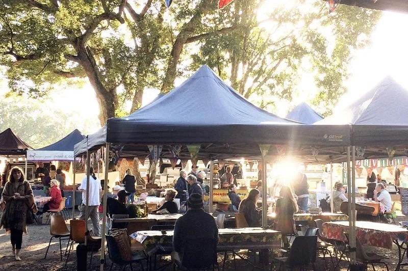 Outdoor market with tents, people sitting and walking, trees in the background, and sunlight filtering through.