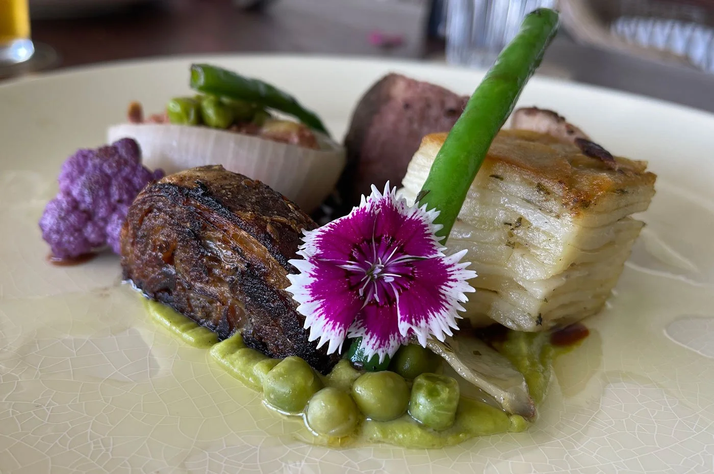 A gourmet dish with layered potatoes, green beans, roasted meat, purple cauliflower, green peas, and garnished with a pink and white edible flower.