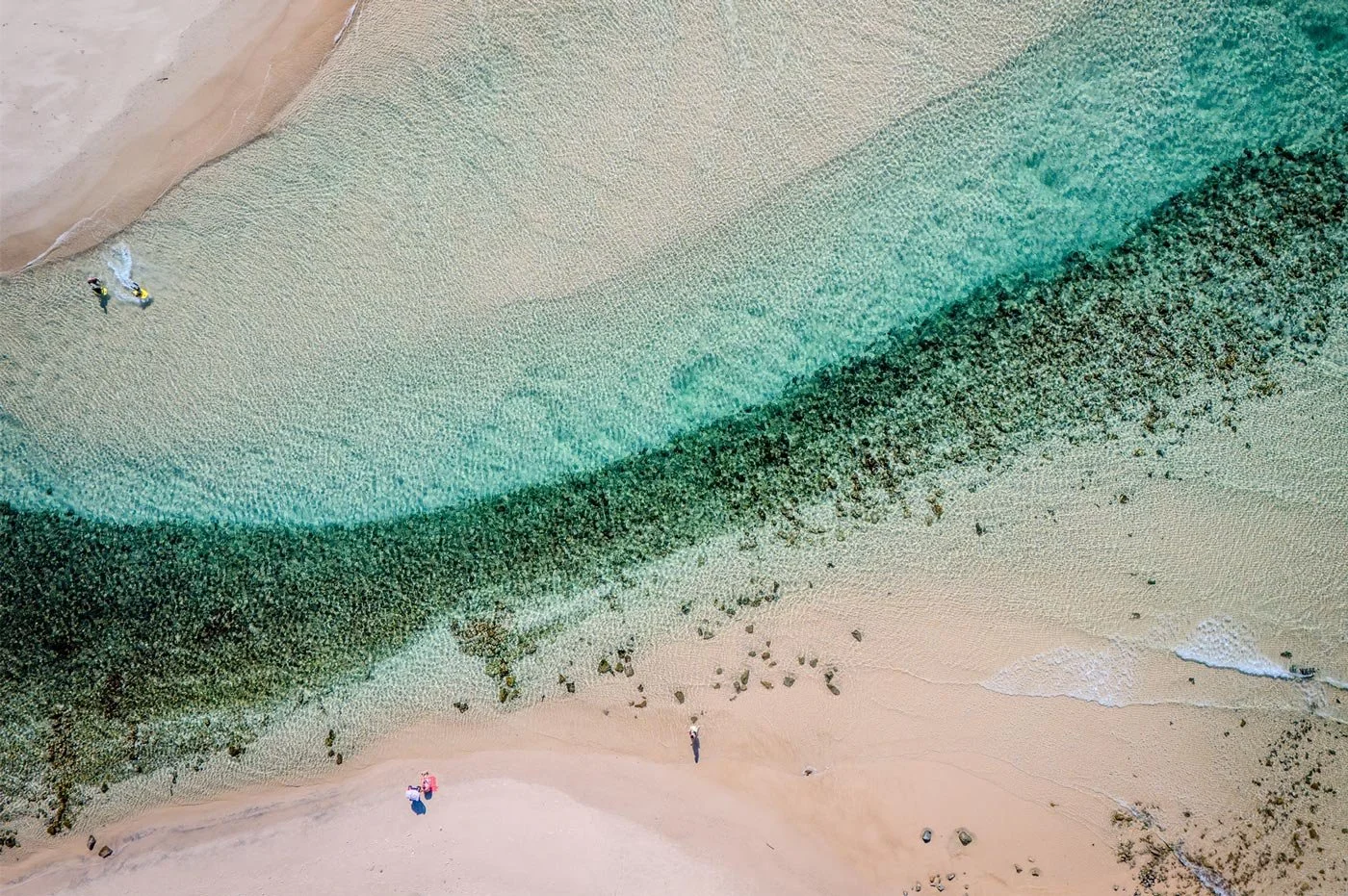 Aerial view of a sandy beach with clear turquoise water, a person standing on the sand, and two people with umbrellas.