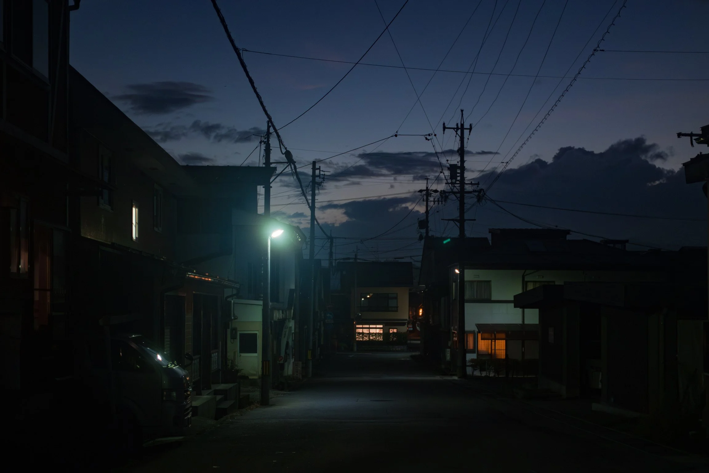 A quiet residential street at dusk with a dark sky, silhouetted buildings, power lines, and a streetlamp casting a soft glow.