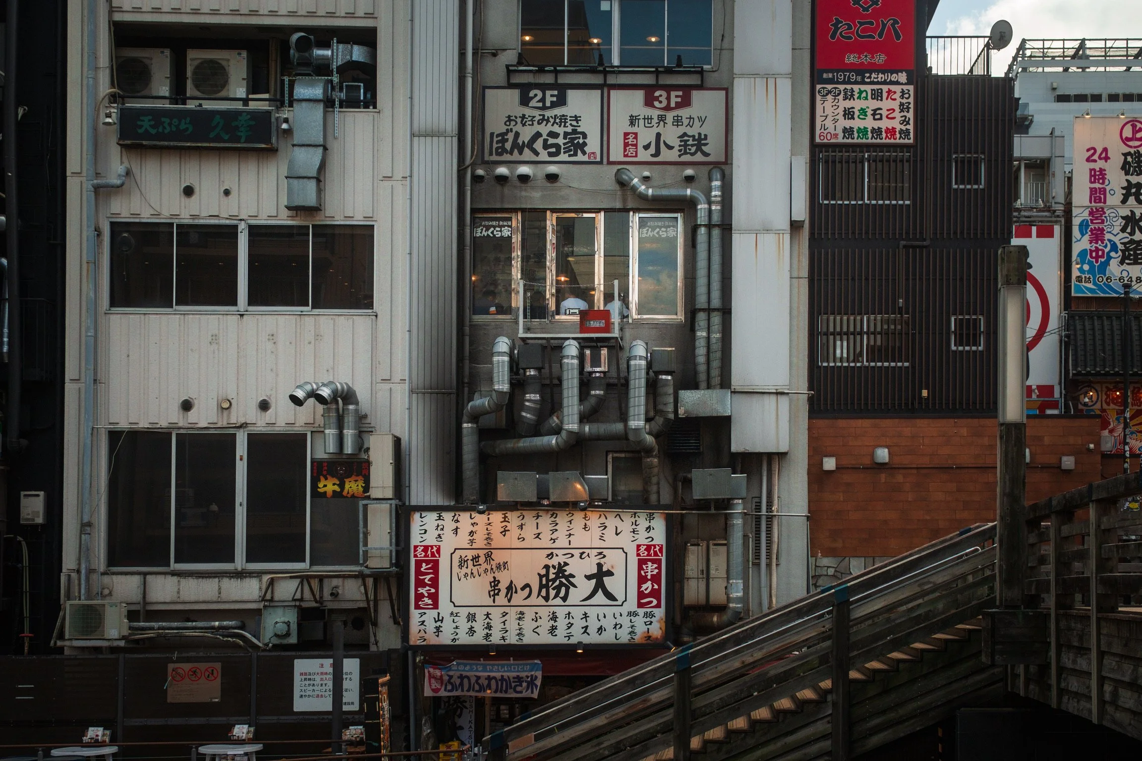 A multi-story building in an urban area with various signs in Japanese, metal ventilation pipes, and air conditioning units on the exterior. There are stairs and small tables at the bottom.