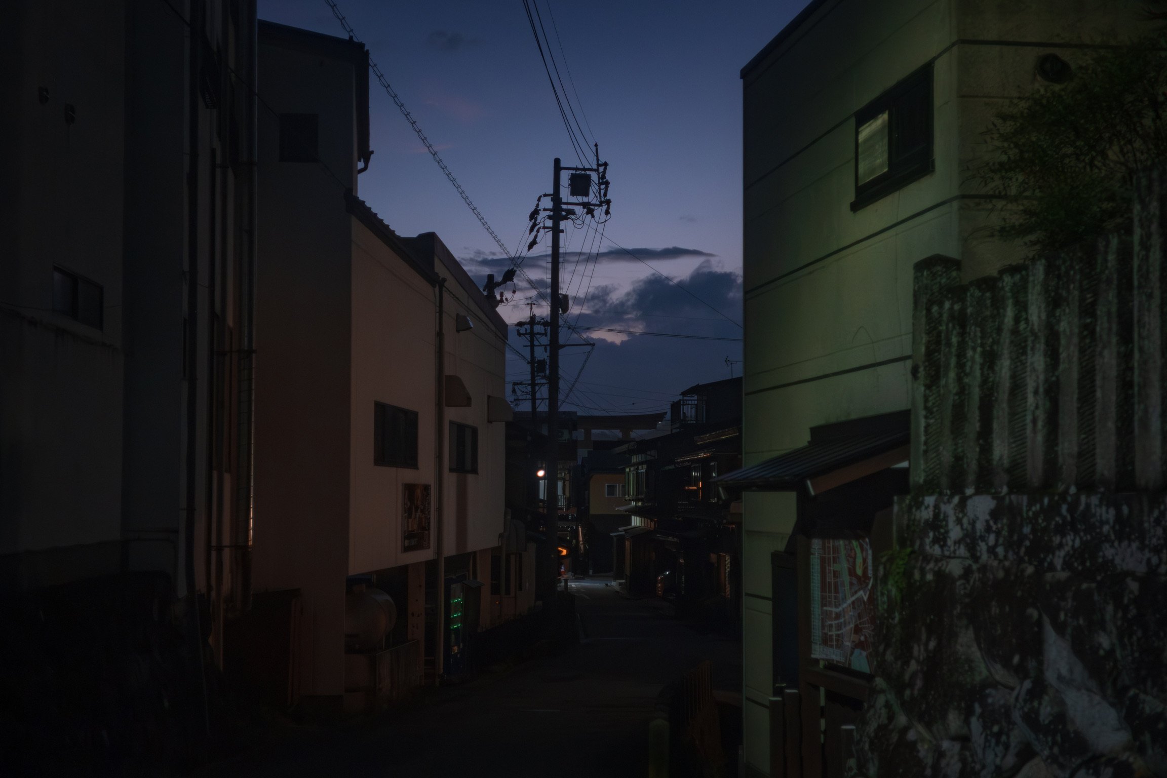 A twilight scene of a residential neighborhood with buildings, power lines, and utility poles under a partly cloudy sky.