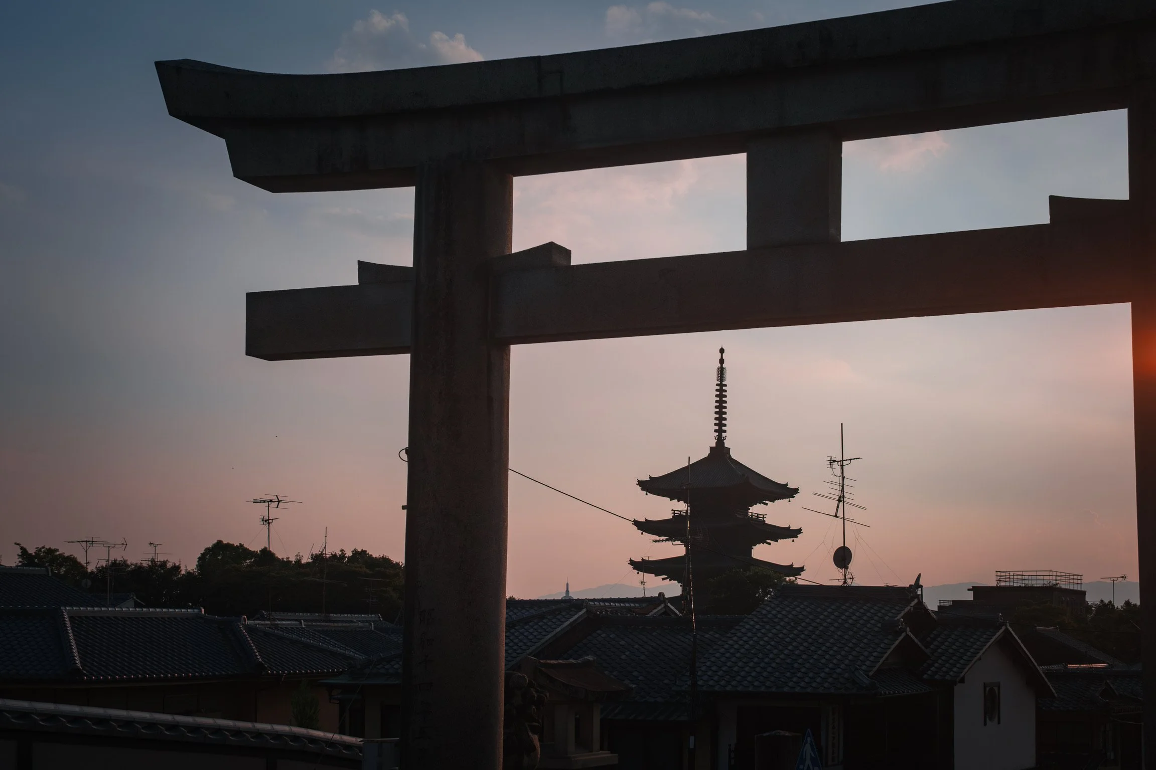 A photo of a traditional Japanese torii gate with a pagoda in the background during sunset, with rooftops and antennas in the foreground.