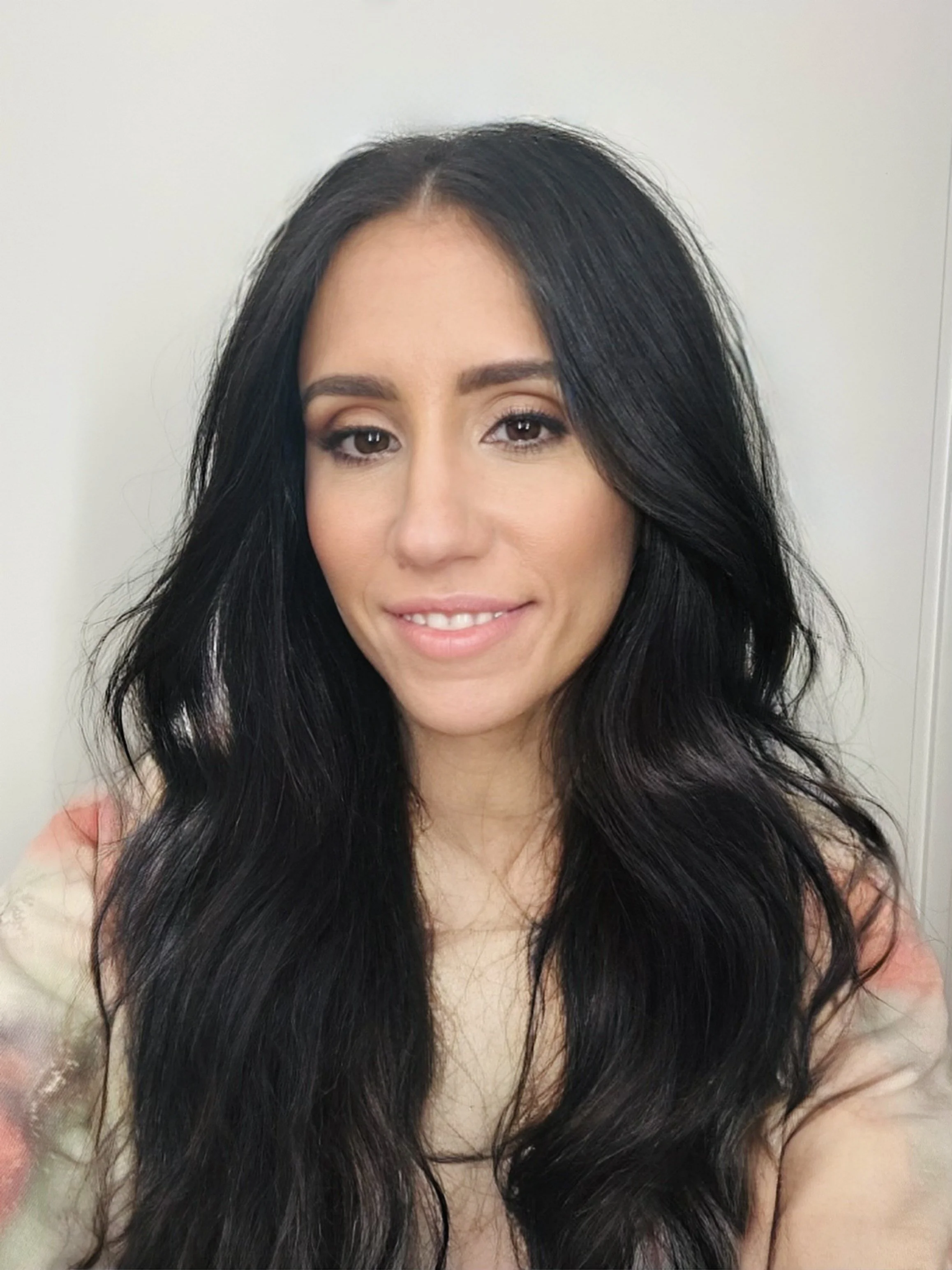 A woman with long black wavy hair, smiling softly, with a light-colored background.
