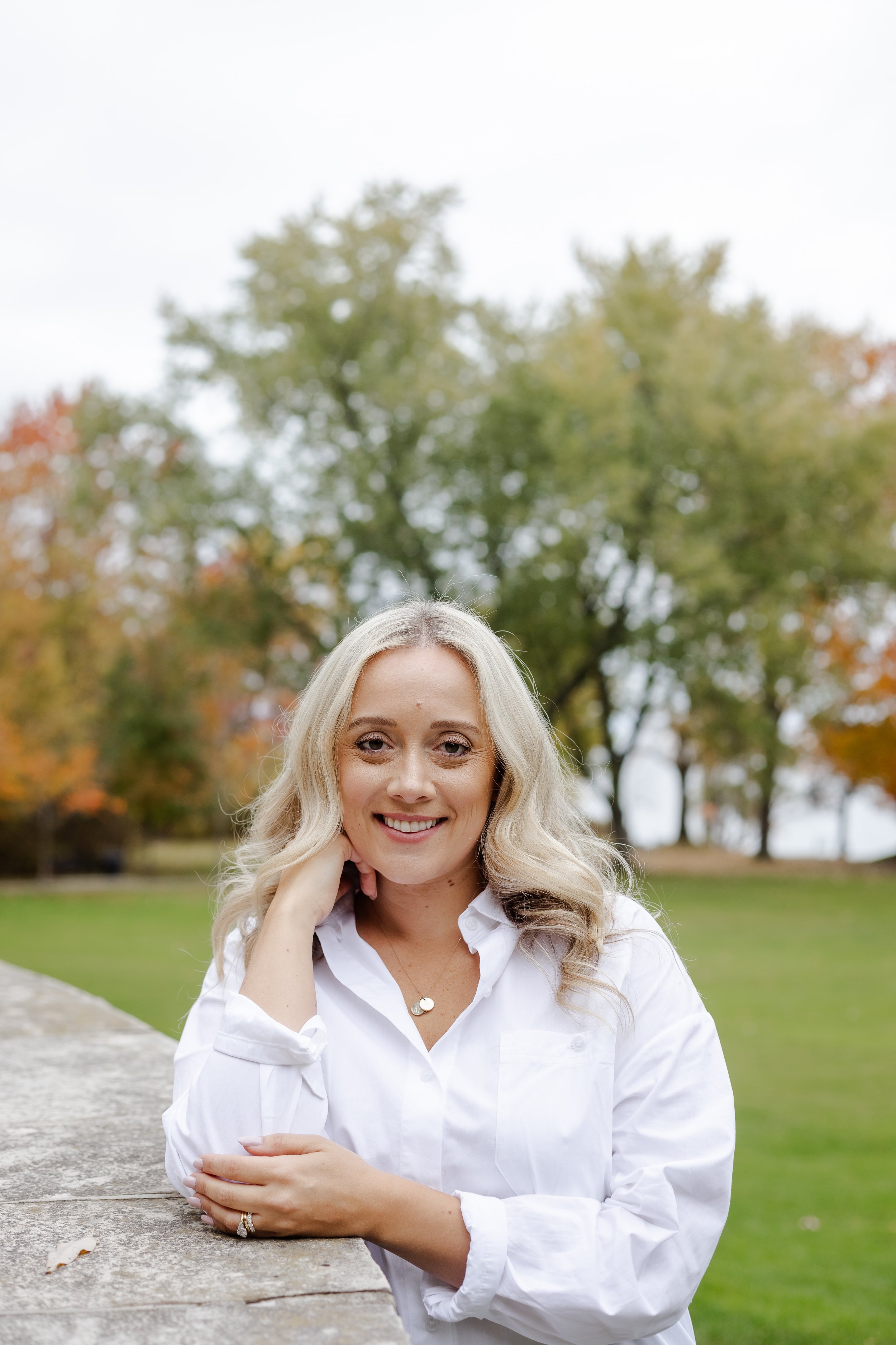 A woman with long blonde hair, wearing a white blouse and a necklace, sitting outdoors near a stone ledge with trees in the background during fall.