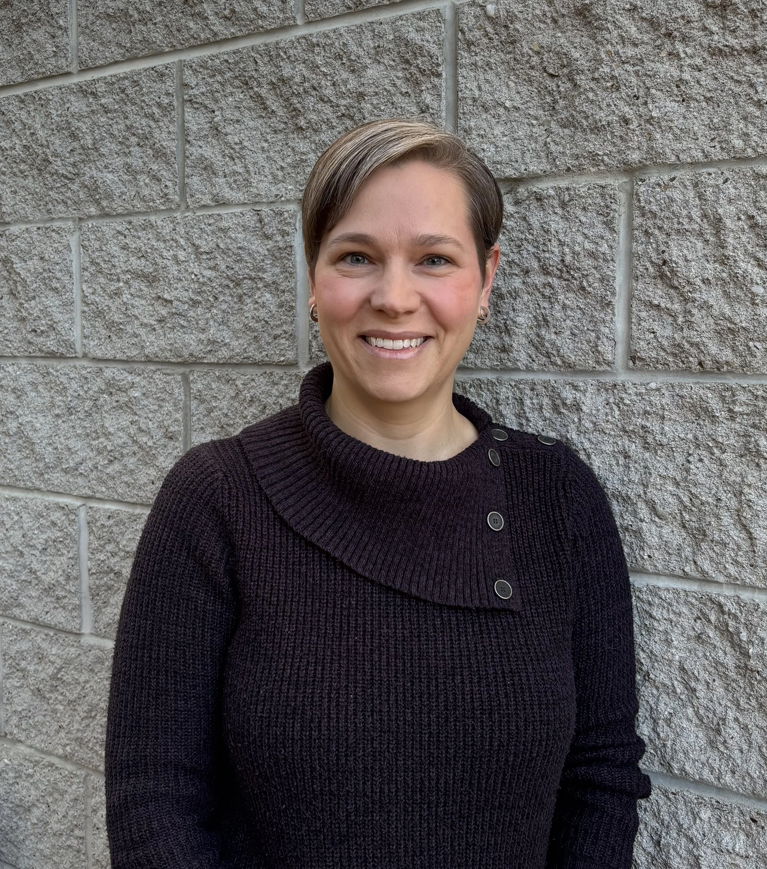 A smiling woman with short hair and earrings wearing a black sweater standing in front of a gray brick wall.