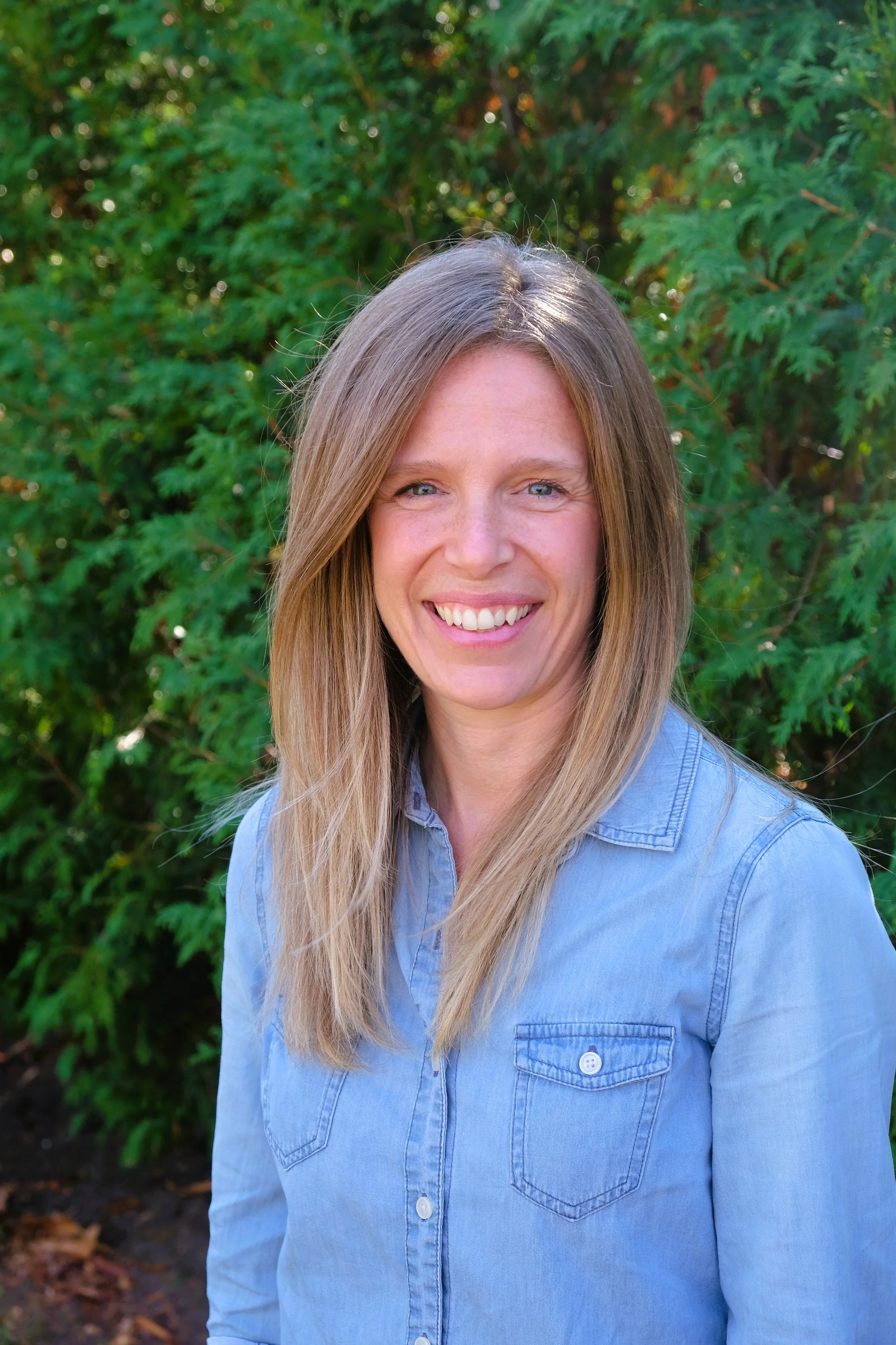 A woman with long blonde hair, smiling, wearing a light blue denim shirt, stands outdoors with green foliage in the background.