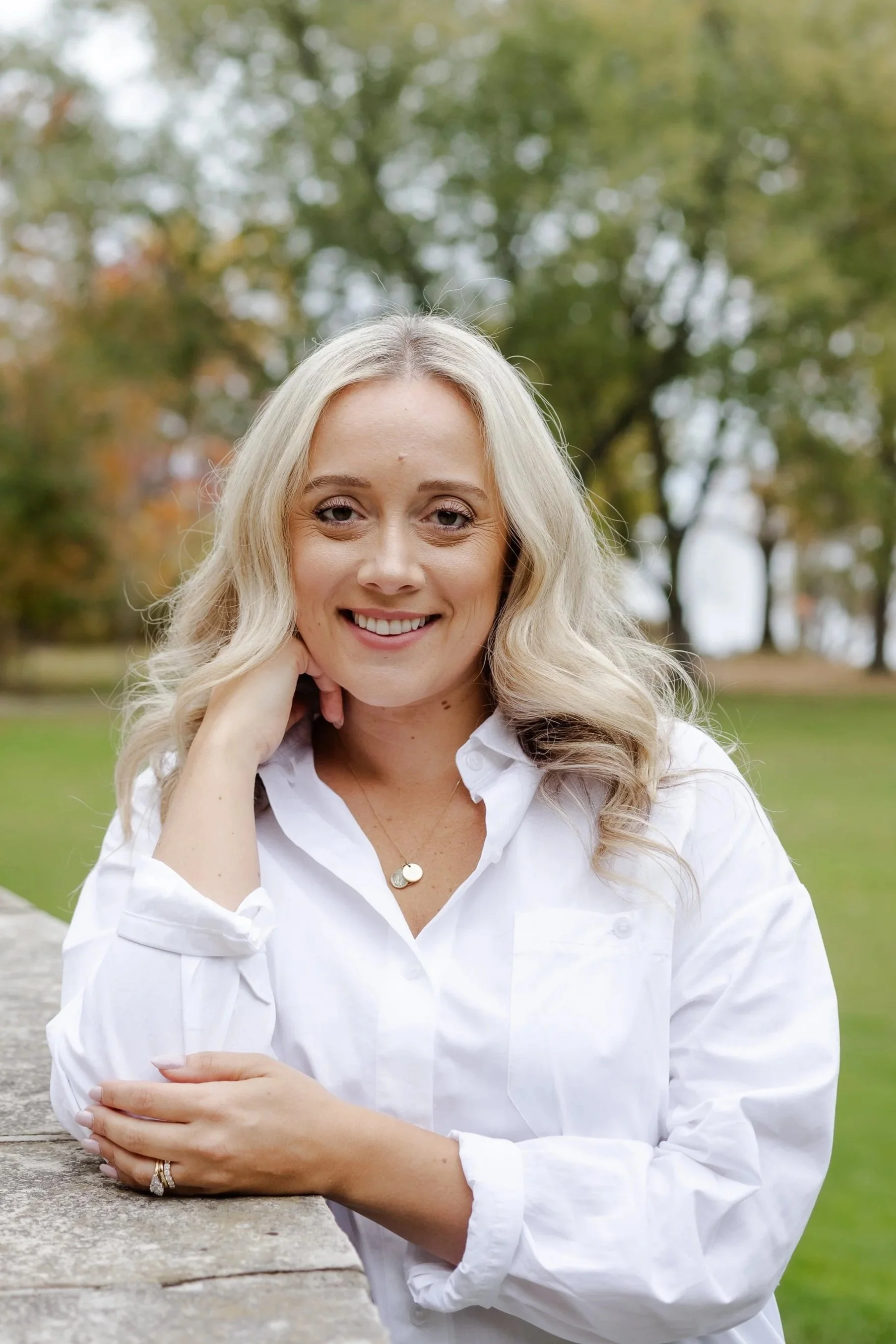 A smiling woman with blonde hair in a white shirt leaning on a stone railing outdoors with trees in the background.