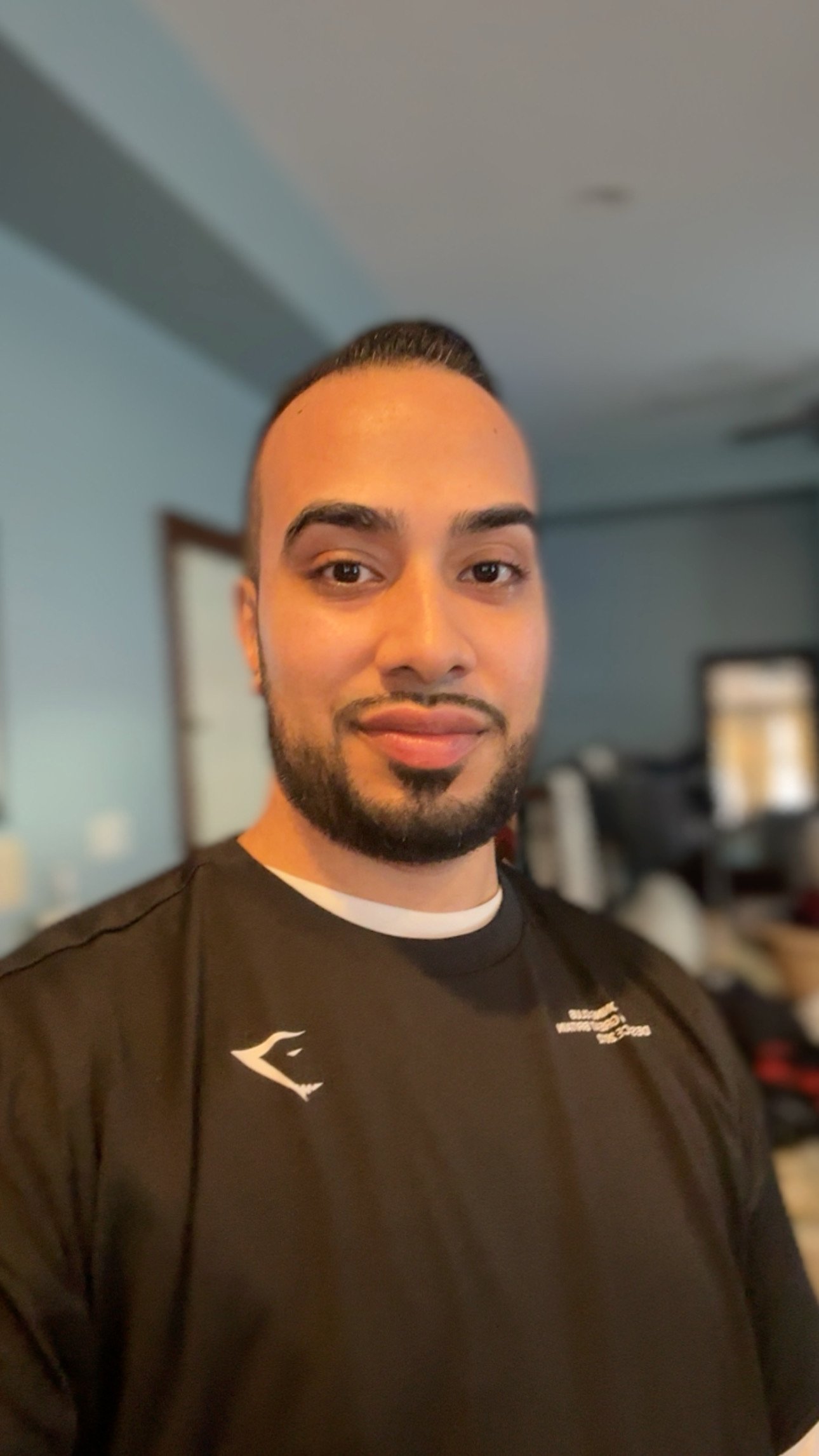 A man smiling with a beard and short dark hair, wearing a black shirt with white details, standing in a room with blue walls and a blurred background.