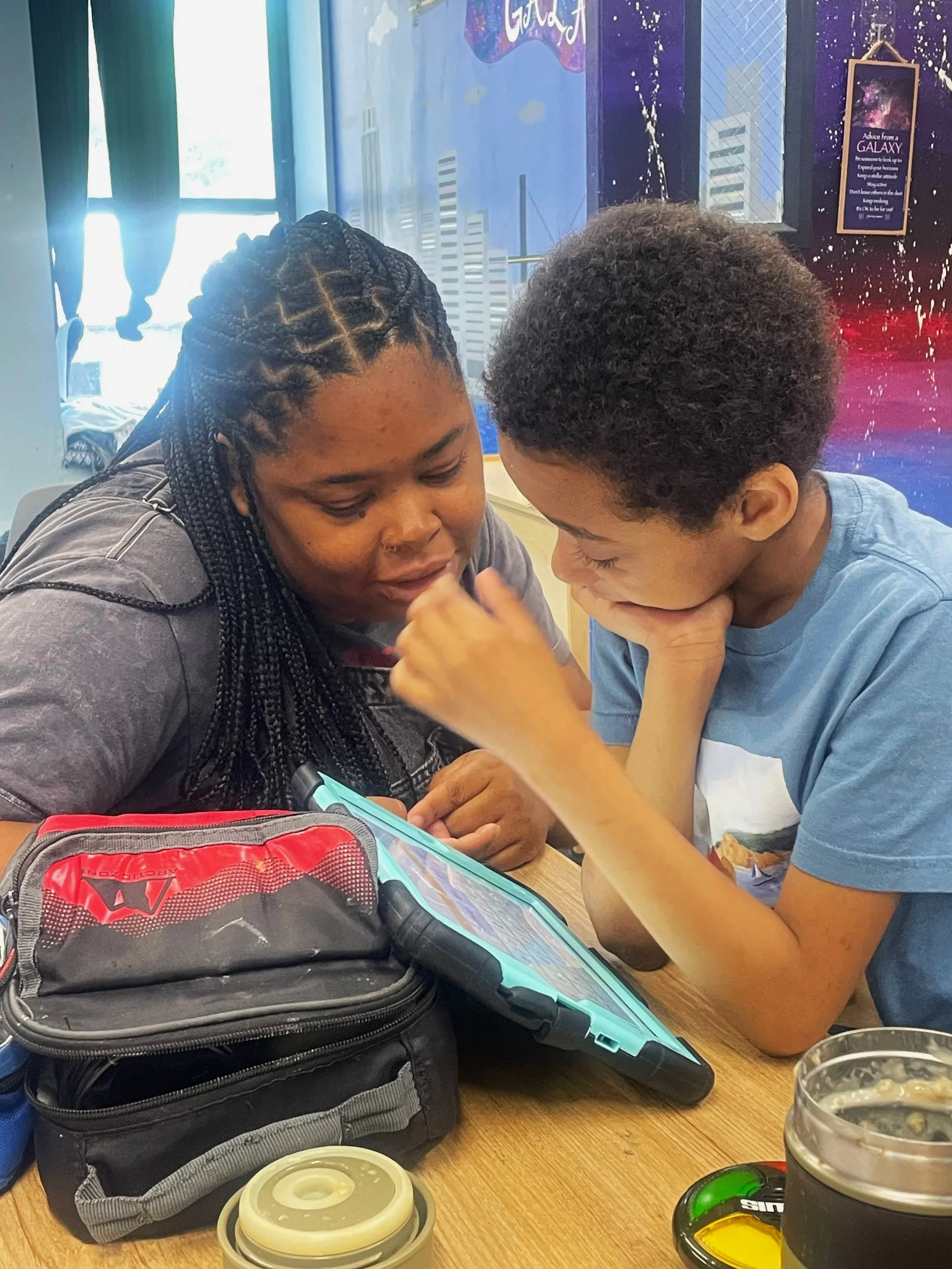 Two children sitting at a table, looking at a tablet device together in a public setting, with backpacks, food containers, and drinks on the table.