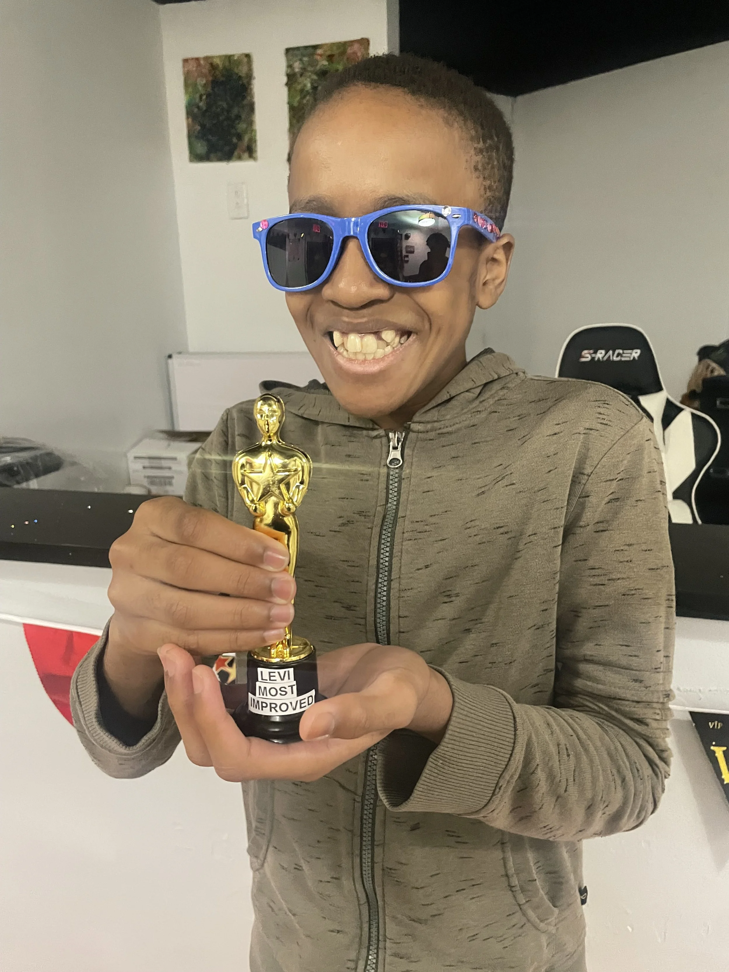 A young man wearing blue sunglasses, smiling, holding a small golden award trophy, with a white wall background and a gaming chair behind him.