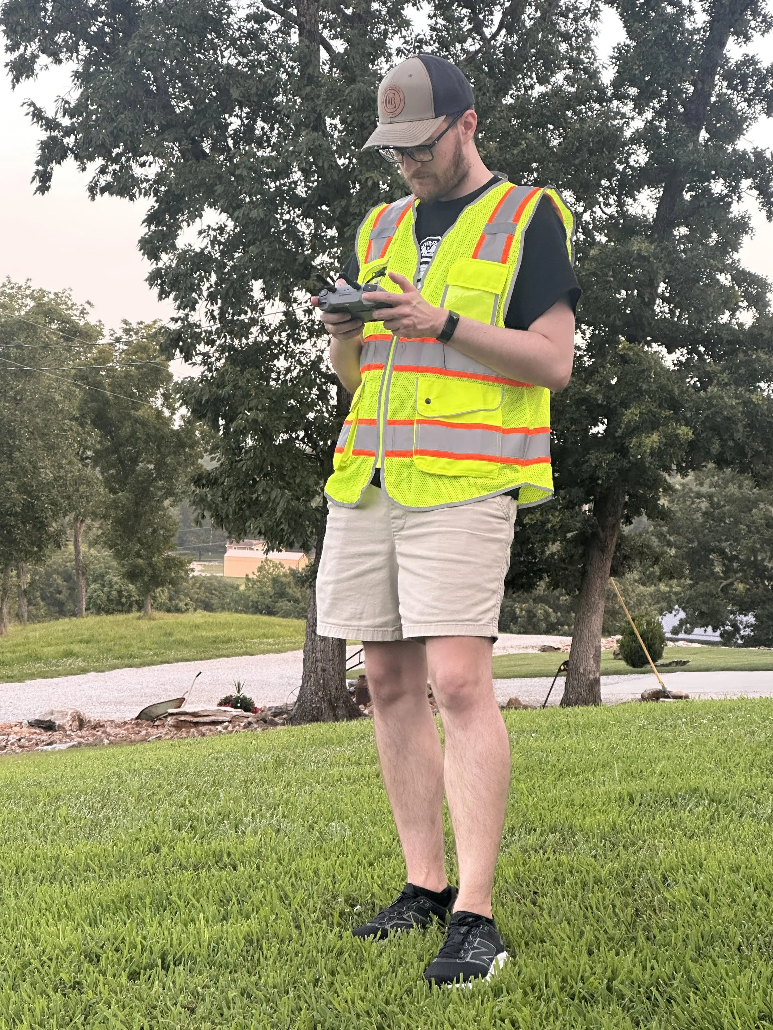 A man standing outdoors on green grass, looking at a device in his hands. He is wearing a black t-shirt, beige shorts, a yellow reflective safety vest, a baseball cap, glasses, and black sneakers.
