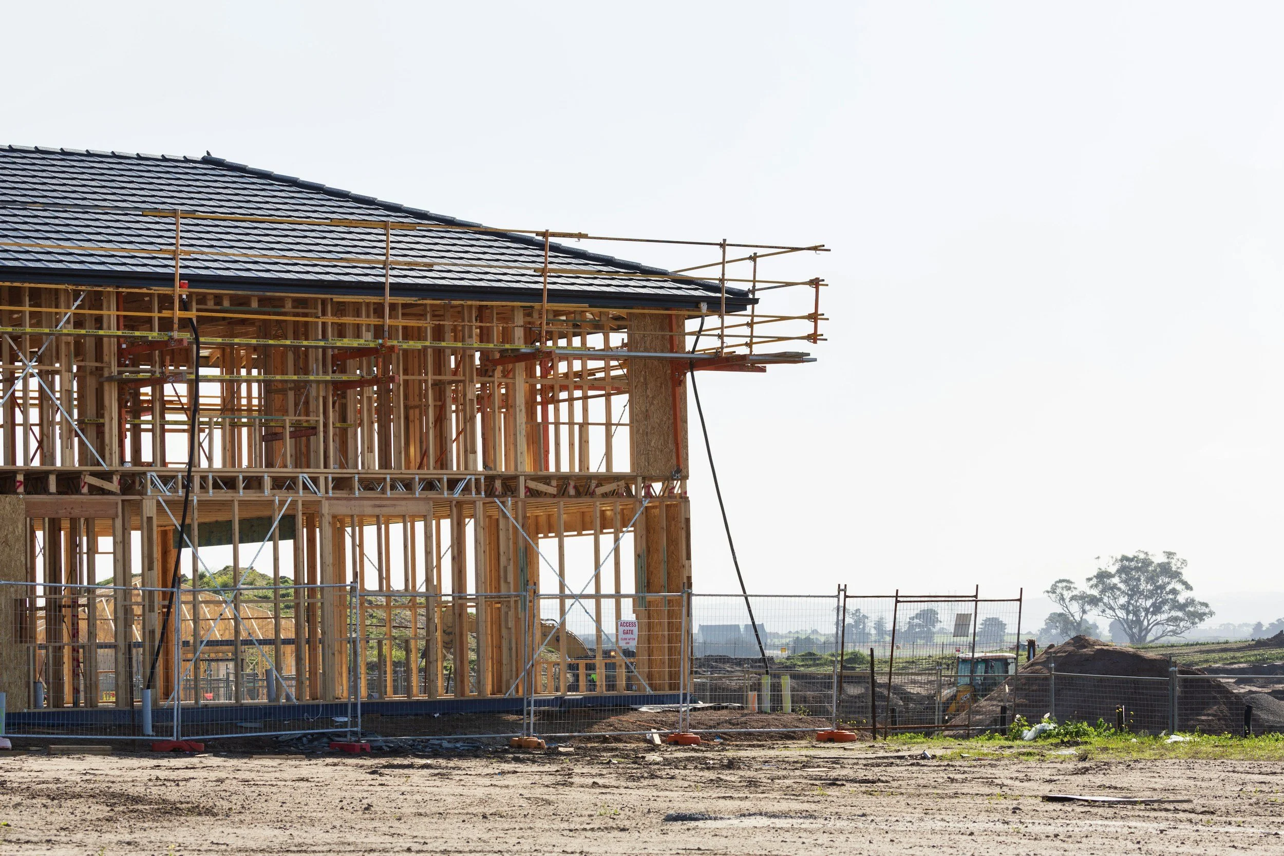 Construction site with wooden framing for a building, scaffolding, and a partly completed roof with shingles, under a clear sky.