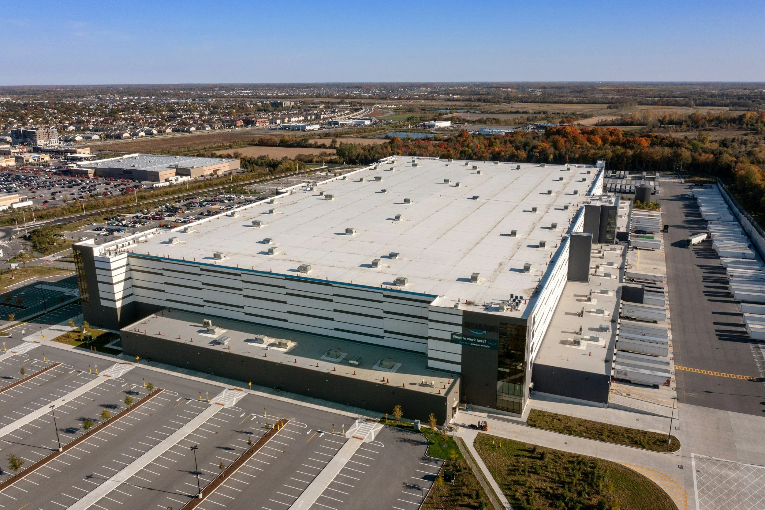 Aerial view of a large Amazon warehouse with surrounding parking lots and an expansive landscape including trees and fields in the background.